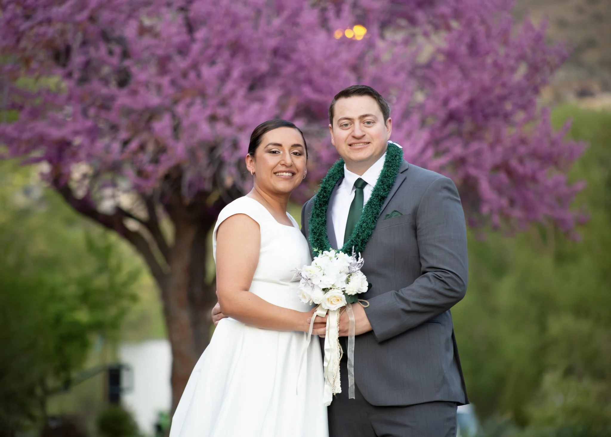 A newlywed couple in wedding attire, standing outdoors in front of a large tree with pink blossoms. The bride has dark hair and is wearing a white dress, holding a bouquet of white flowers. The groom has short dark hair, wearing a gray suit, white sh