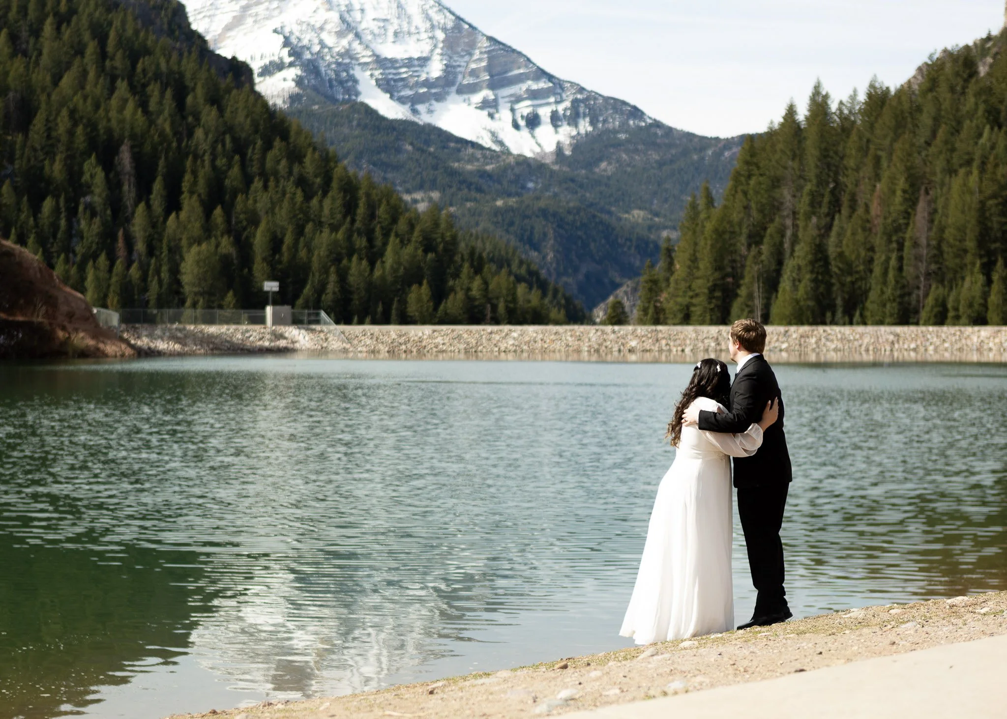 A bride and groom embracing on the edge of a lake in a mountainous region, with snow-capped peaks and dense pine forests in the background.