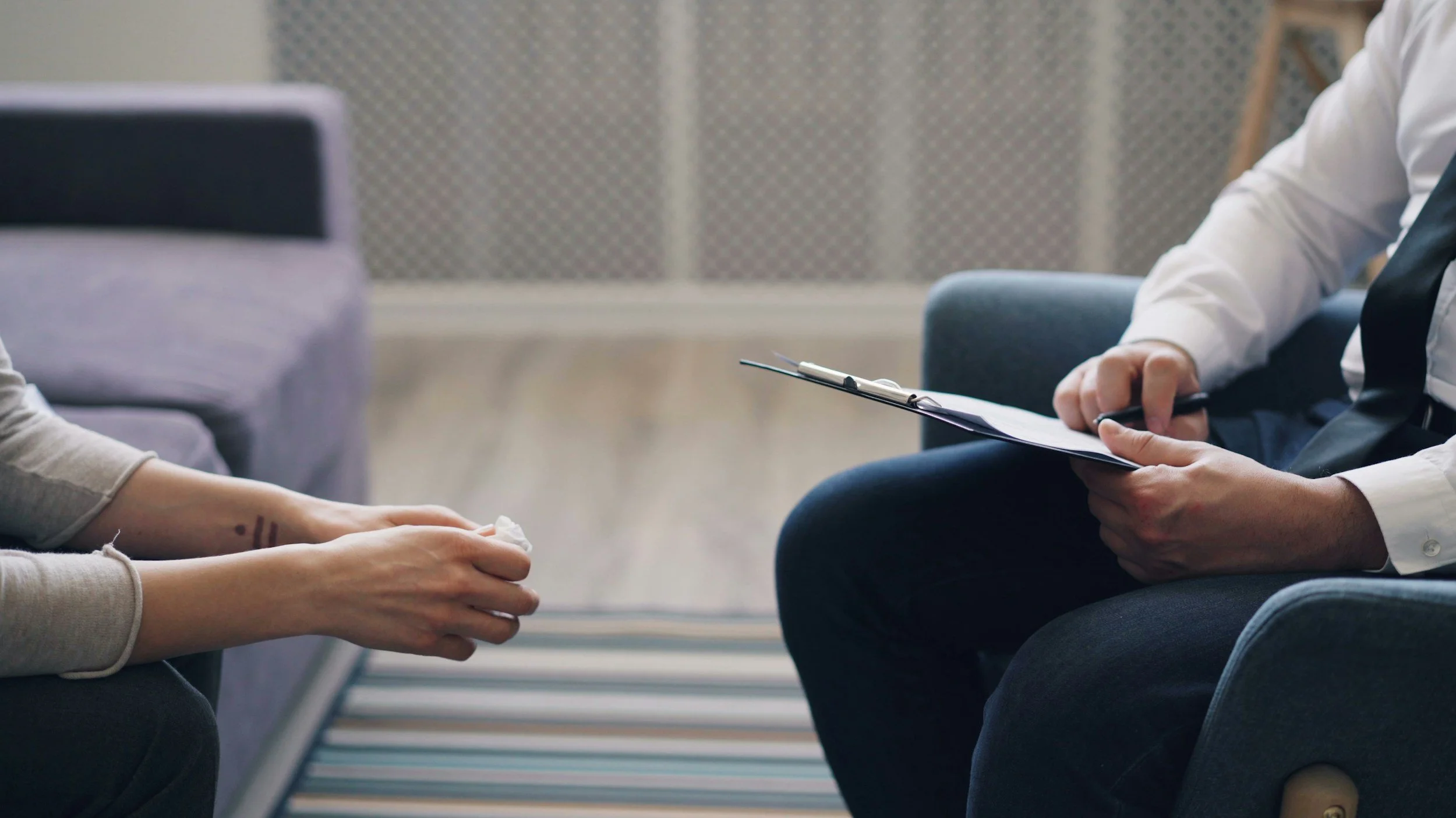 Person sitting in a therapy or counseling session, holding their hands out, with a therapist taking notes on a clipboard.