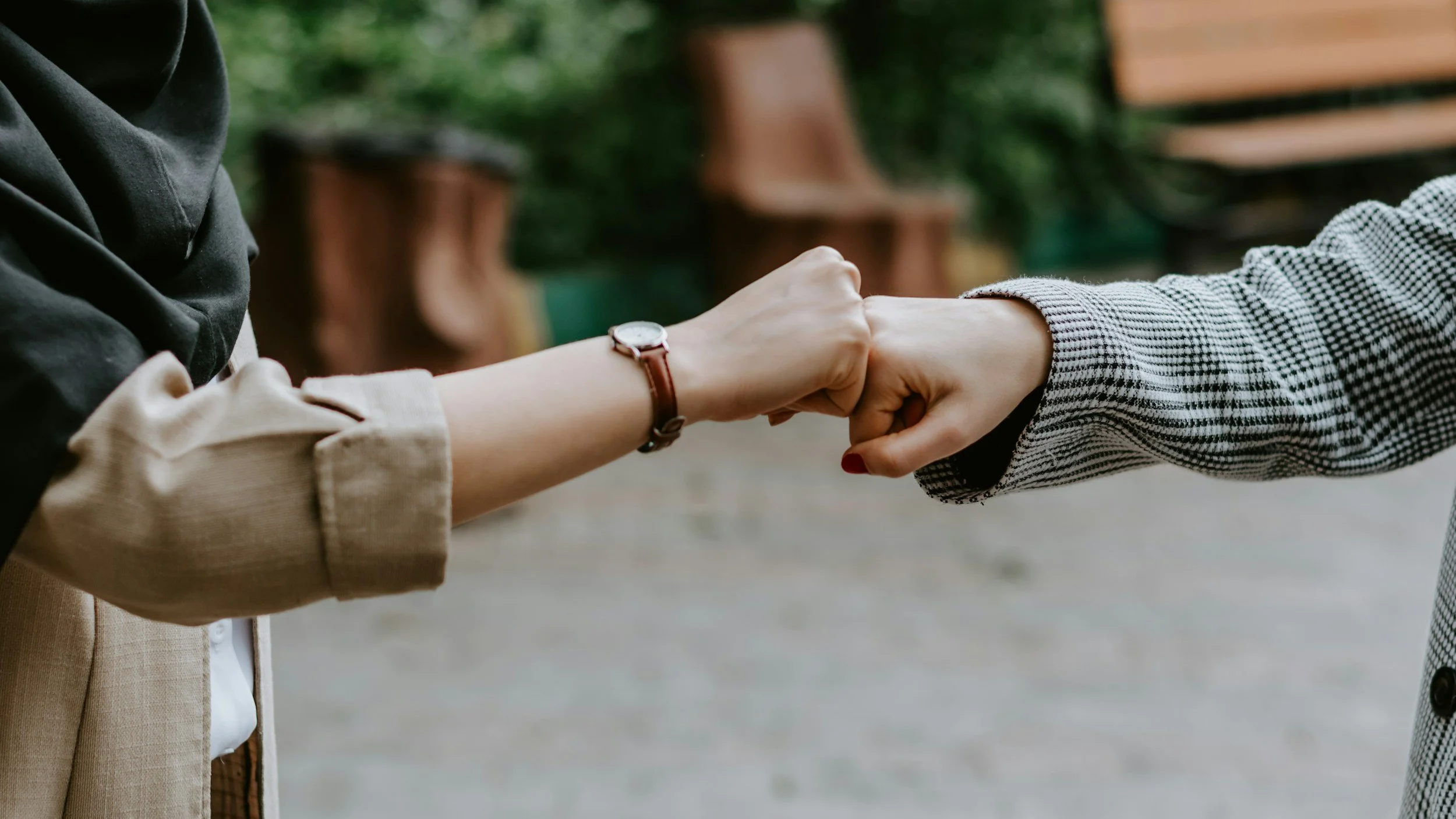 Two people fist bumping outdoors with blurred greenery and benches in the background.