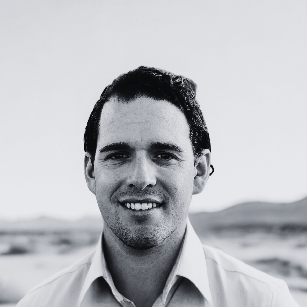 Black-and-white photo of a smiling man with dark, wavy hair, wearing a collared shirt, outdoors with a blurred landscape in the background.