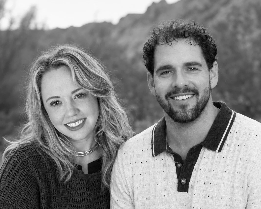 A black-and-white photo of a smiling woman with wavy hair and a woman with curly hair, both wearing casual tops, outdoors with mountains in the background.