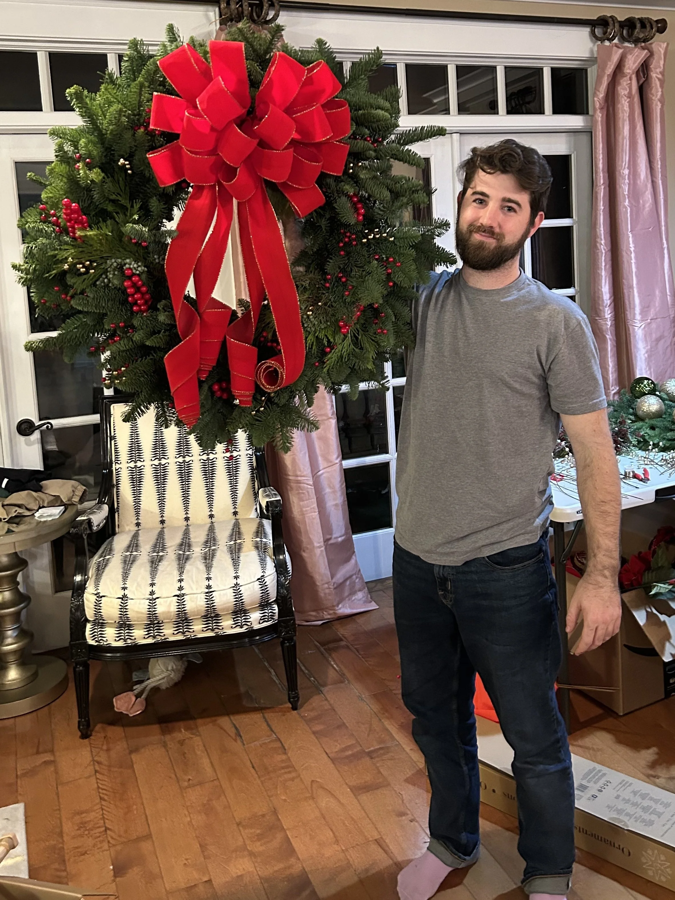 A man with a beard and curly hair standing next to a decorated Christmas wreath with a big red bow, inside a home with a large window, pink curtains, and holiday decorations surrounding him.