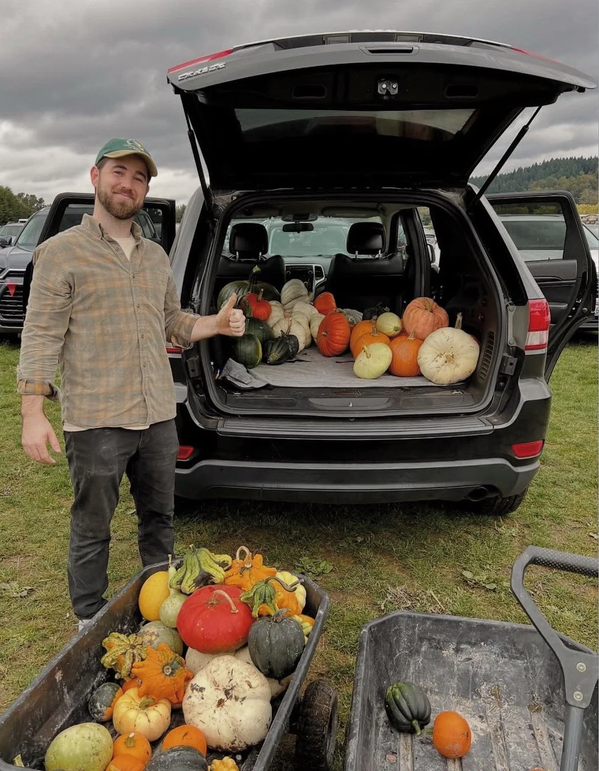 A man standing next to an open car trunk filled with various pumpkins and gourds. There are additional pumpkins and gourds in carts on the ground, and he is giving a thumbs-up.
