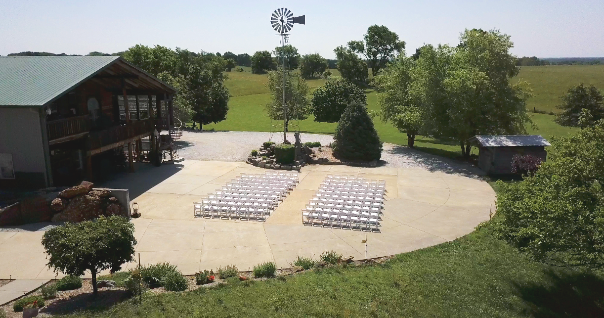 Drone at a wedding Outdoor venue with rows of white chairs arranged on a concrete patio, a windmill, trees, a small shed, and a building with a balcony in the background, set in a rural area.