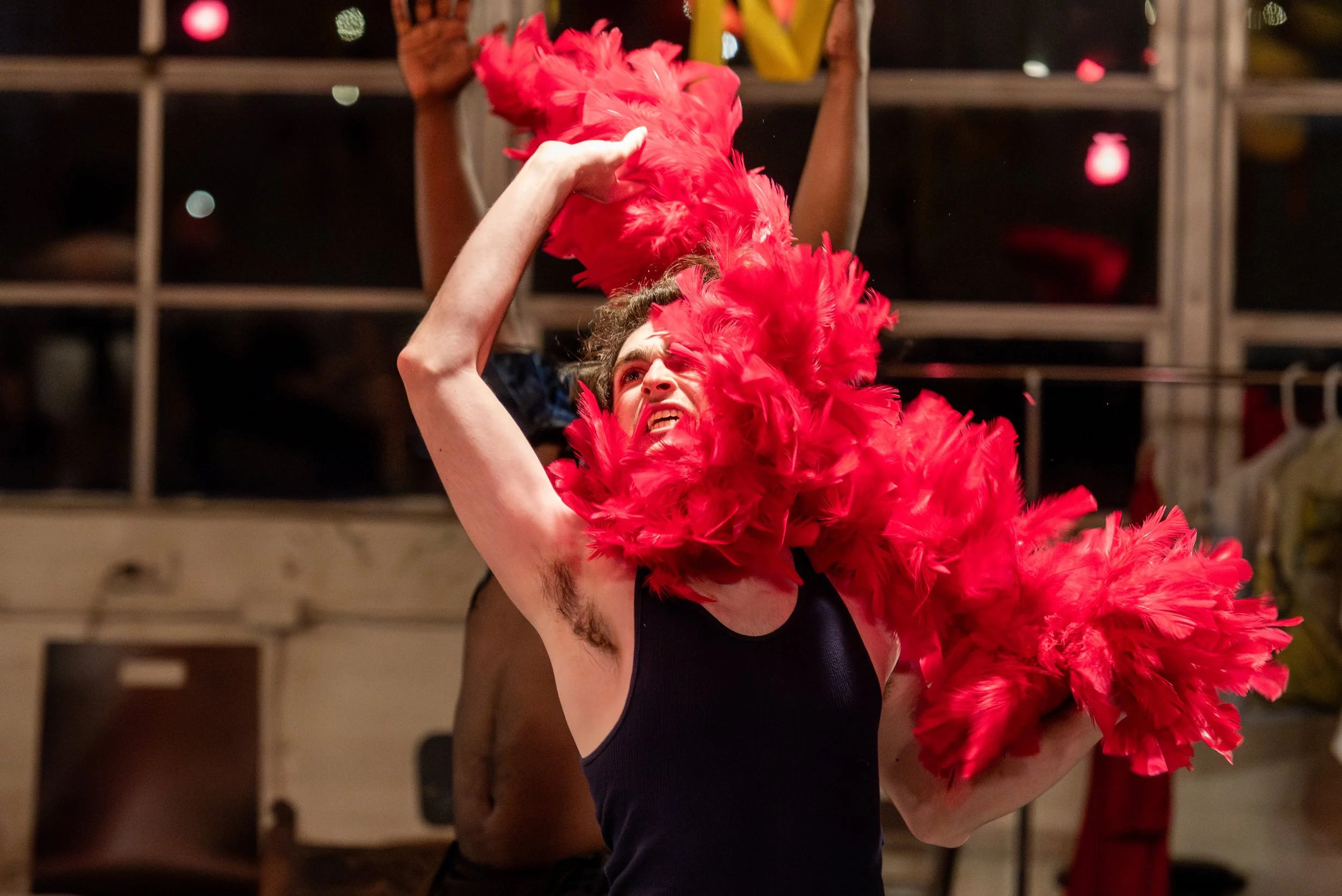 A person wearing a black tank top is adjusting a large red feather boa around their neck and head. They have a playful or expressive facial expression, with some hair visible. The background shows a dimly lit space with wooden shelves and a person be