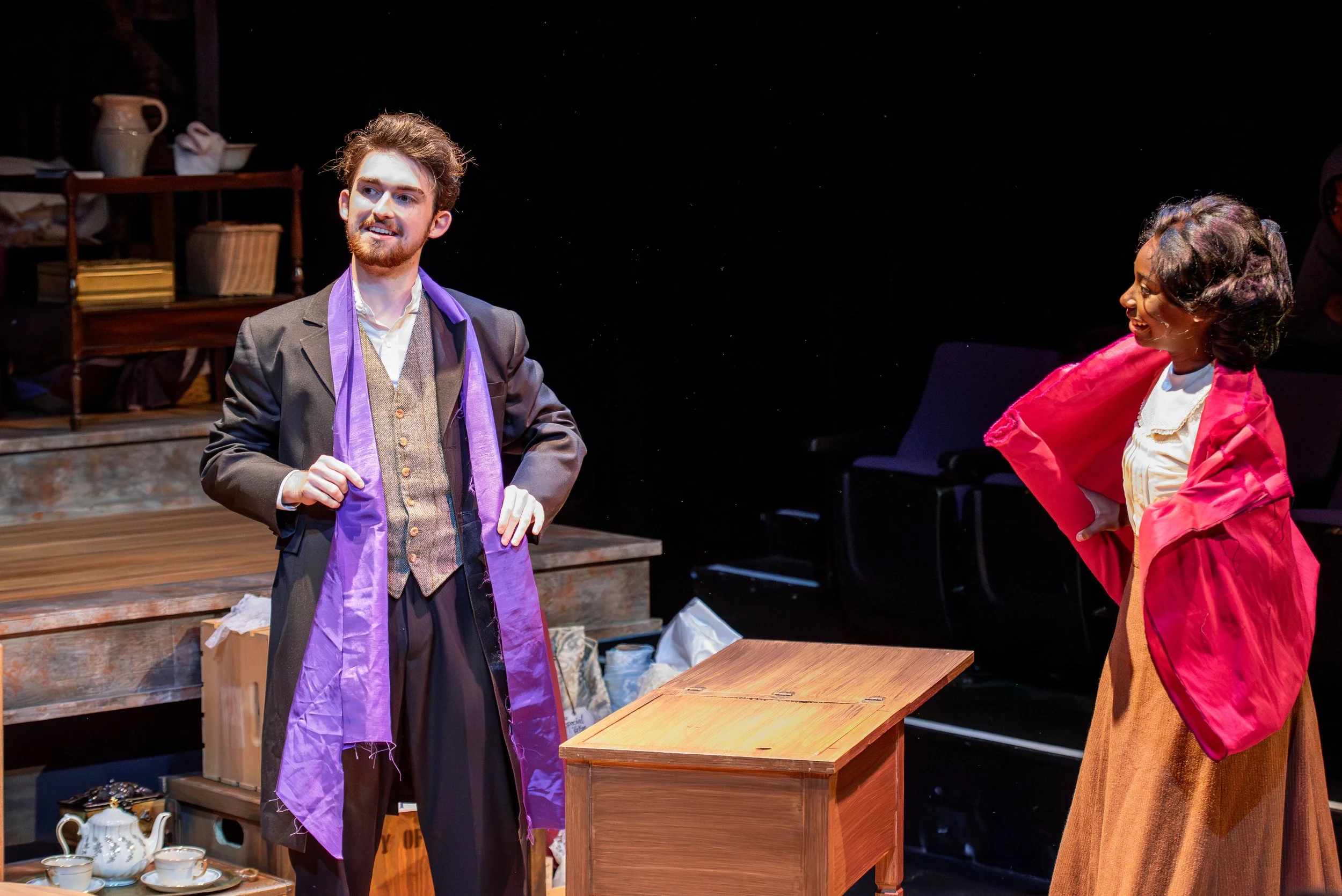 Two actors performing on stage; a man with a beard in a period costume with a purple scarf, and a woman with dark hair in a red jacket and long brown skirt, engaging in a theatrical scene.