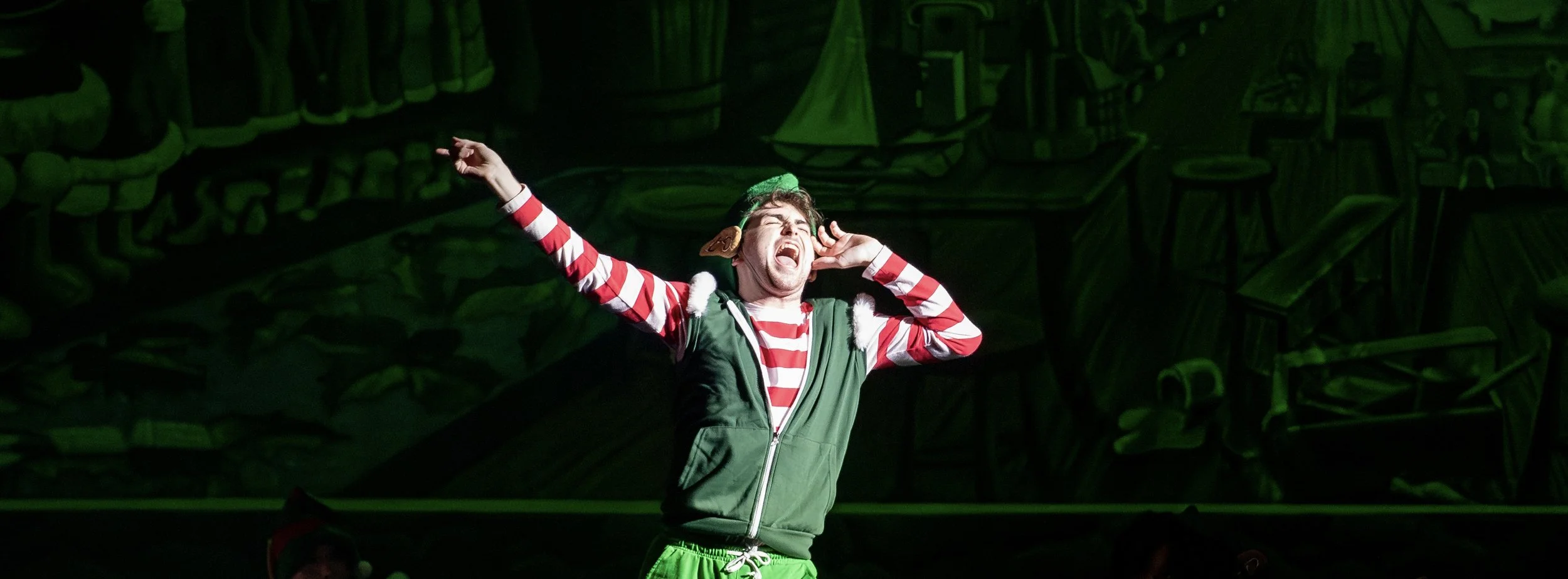 Performer dressed as an elf, singing on stage with expressive gesture, wearing a red and white striped shirt, green vest, and elf accessories, against a dark, patterned background.