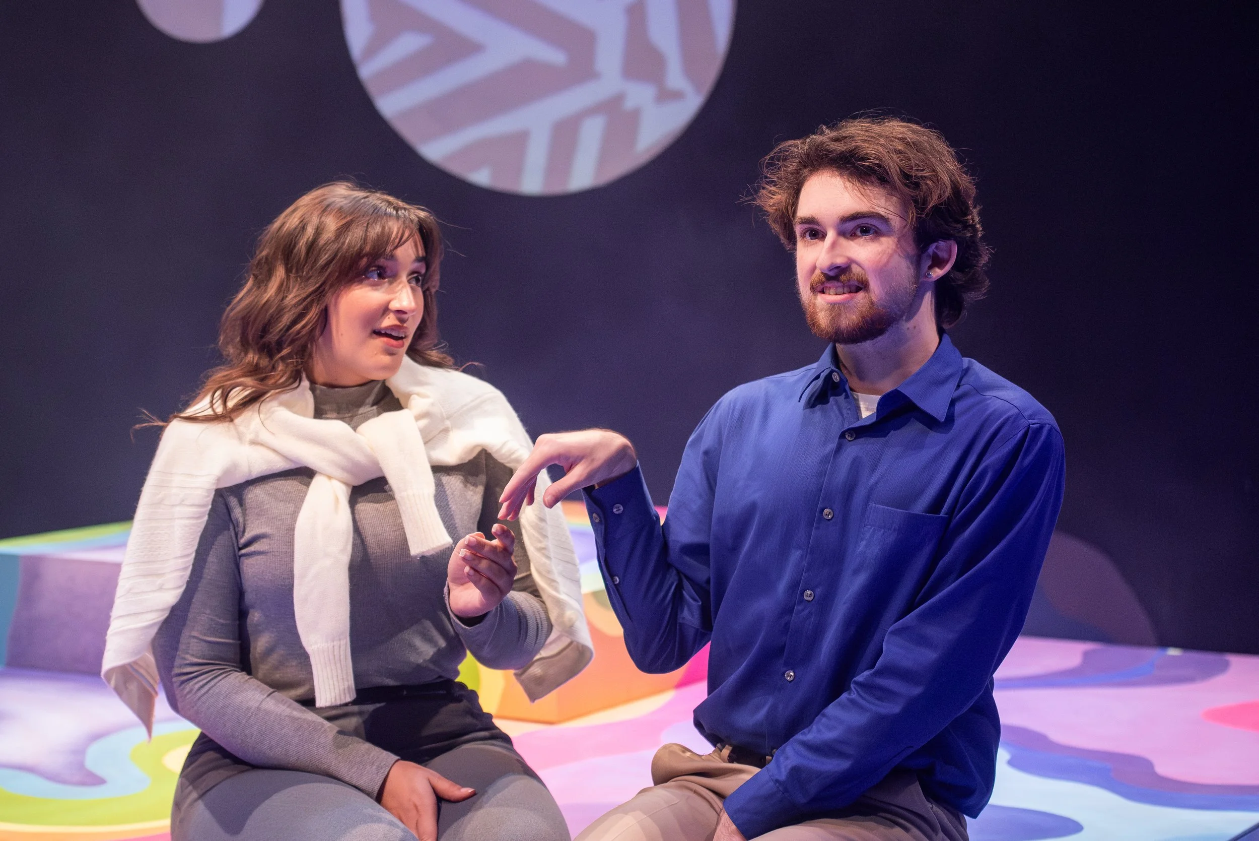 A man and woman are sitting on stage, engaging in a conversation. The woman appears surprised or interested, while the man is speaking and gesturing with his hand. The background features a large circular design.