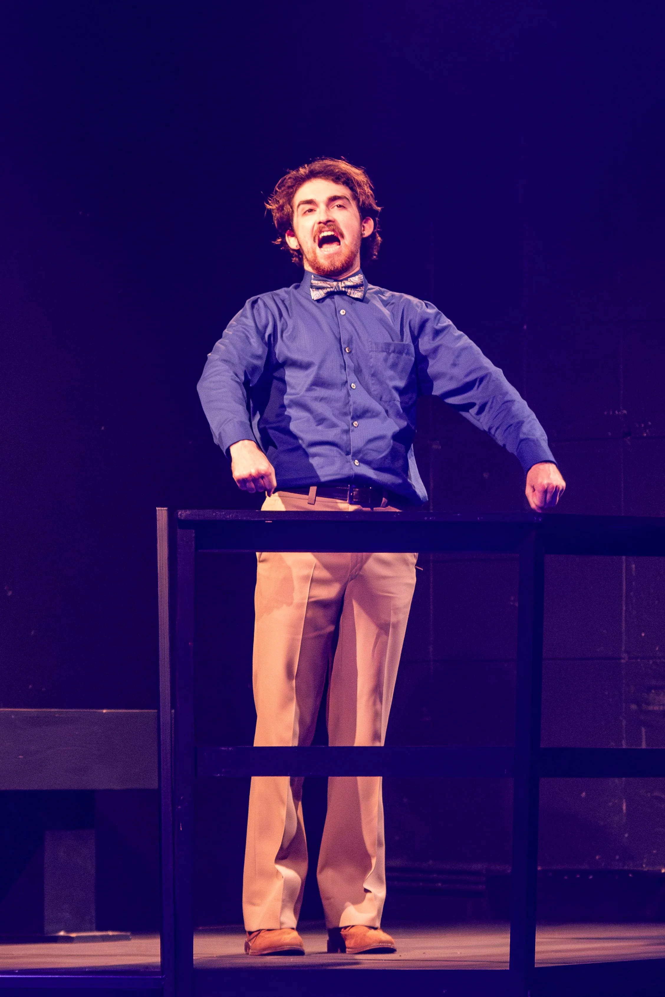 Man with curly hair and beard wearing blue shirt and bow tie performing on stage with a black table in front of him.