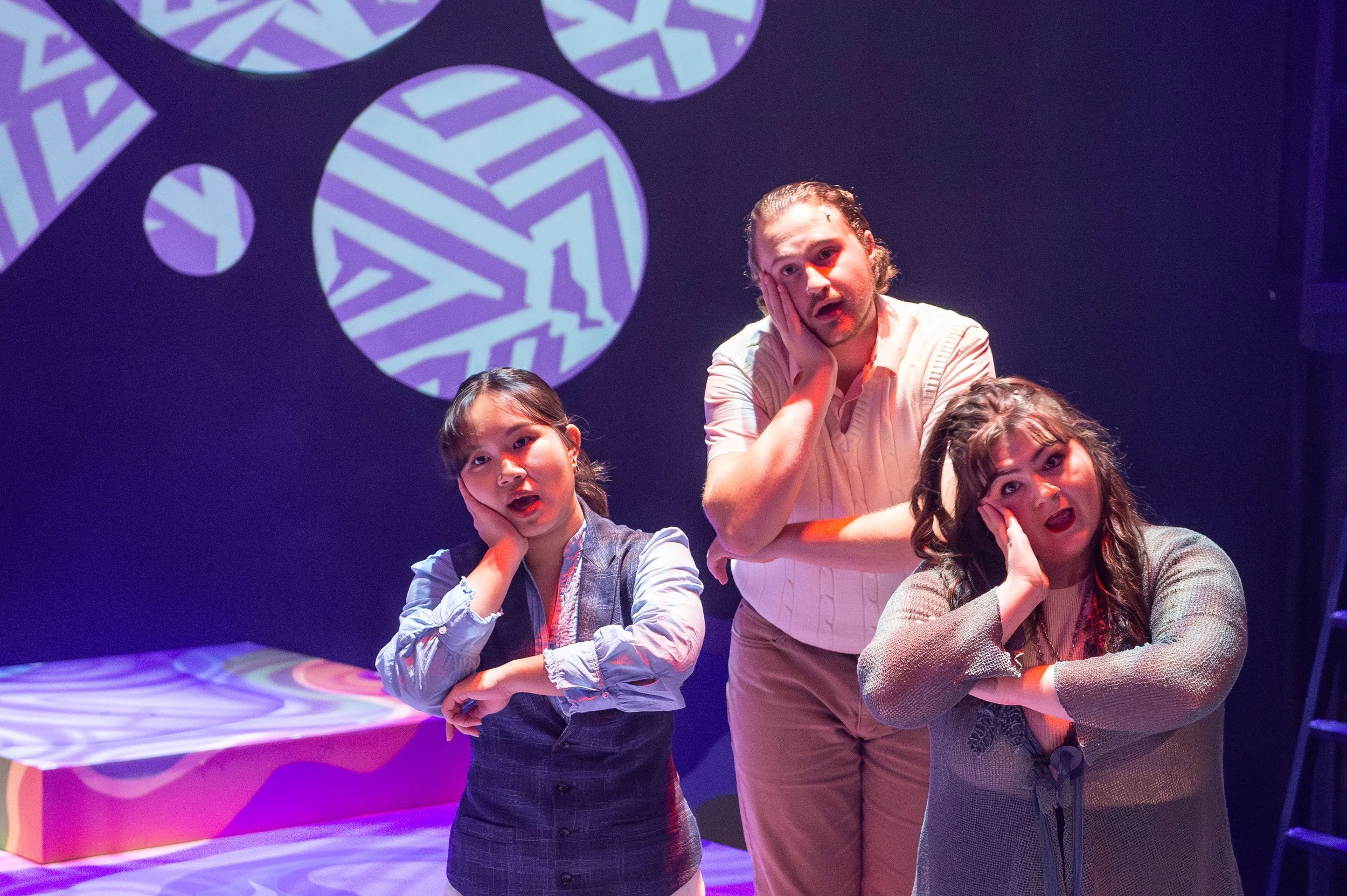 Three people on stage with colorful geometric projections behind them, all holding their faces in surprise or confusion.