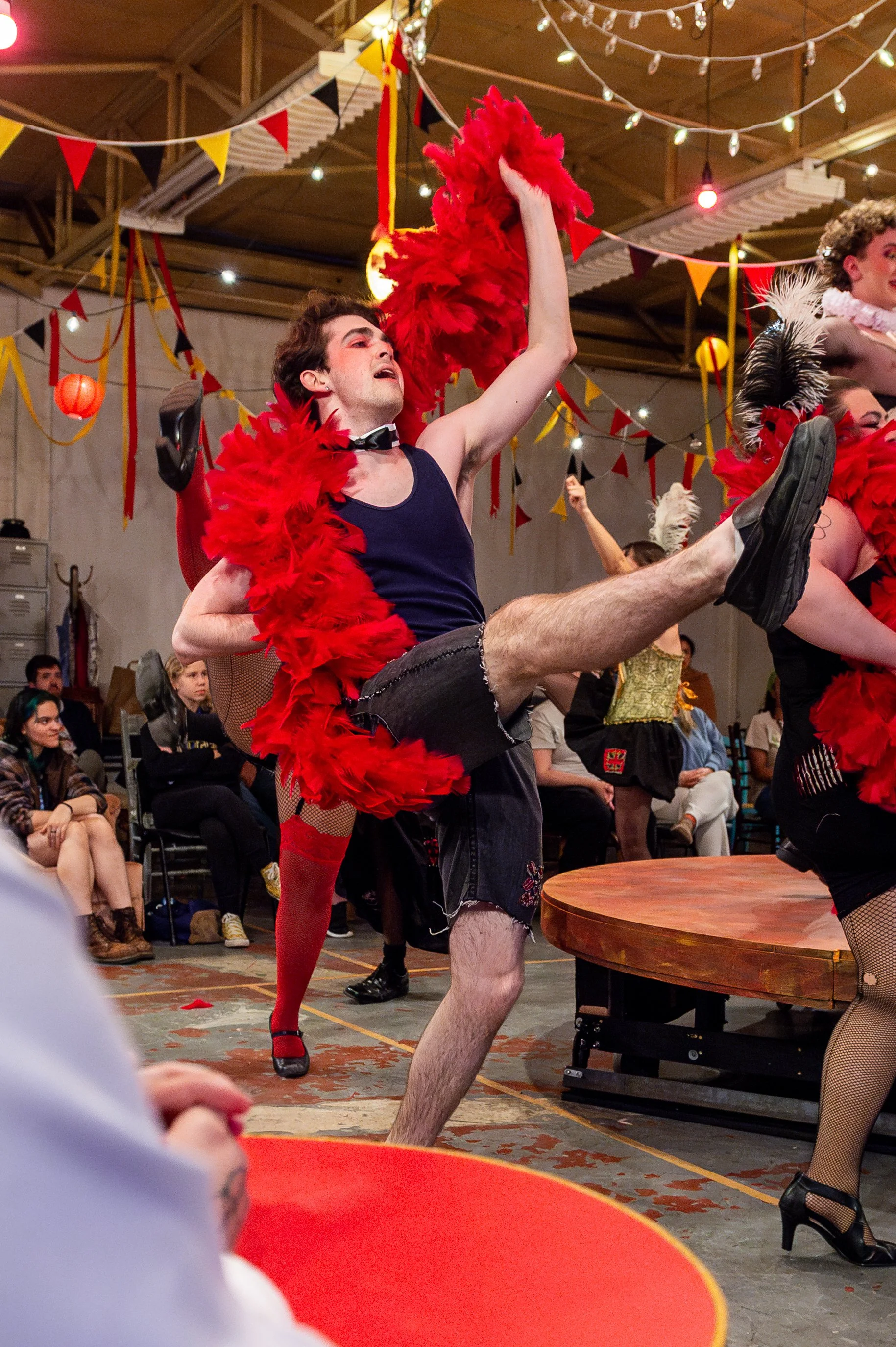Performer in a black tank top, black shorts, and red feather boa dancing with others in a lively, decorated indoor setting.