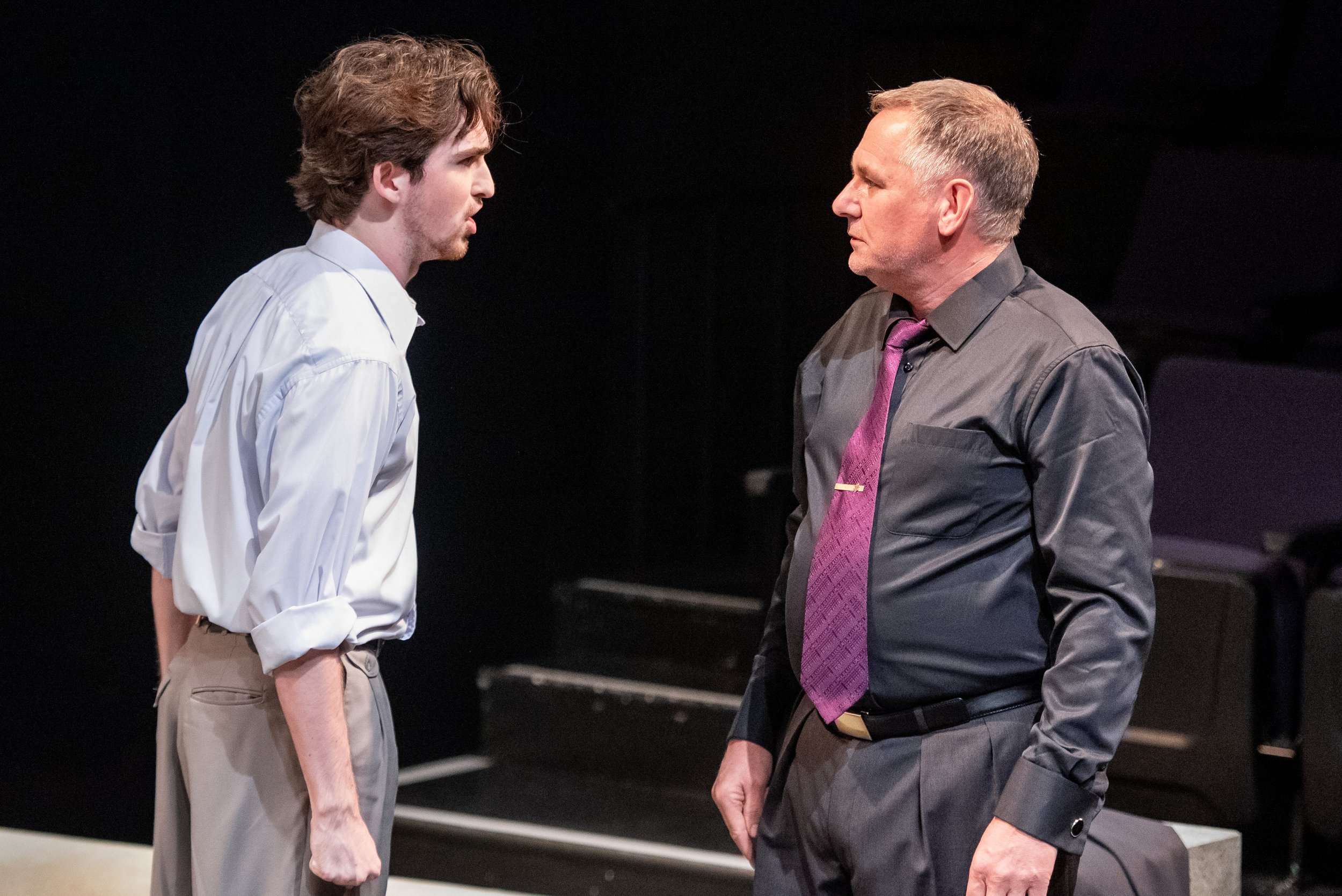 Two men standing face-to-face in a theatrical performance, engaged in an intense conversation. The younger man on the left has curly brown hair and wears a white dress shirt with rolled-up sleeves and beige pants. The older man on the right has short