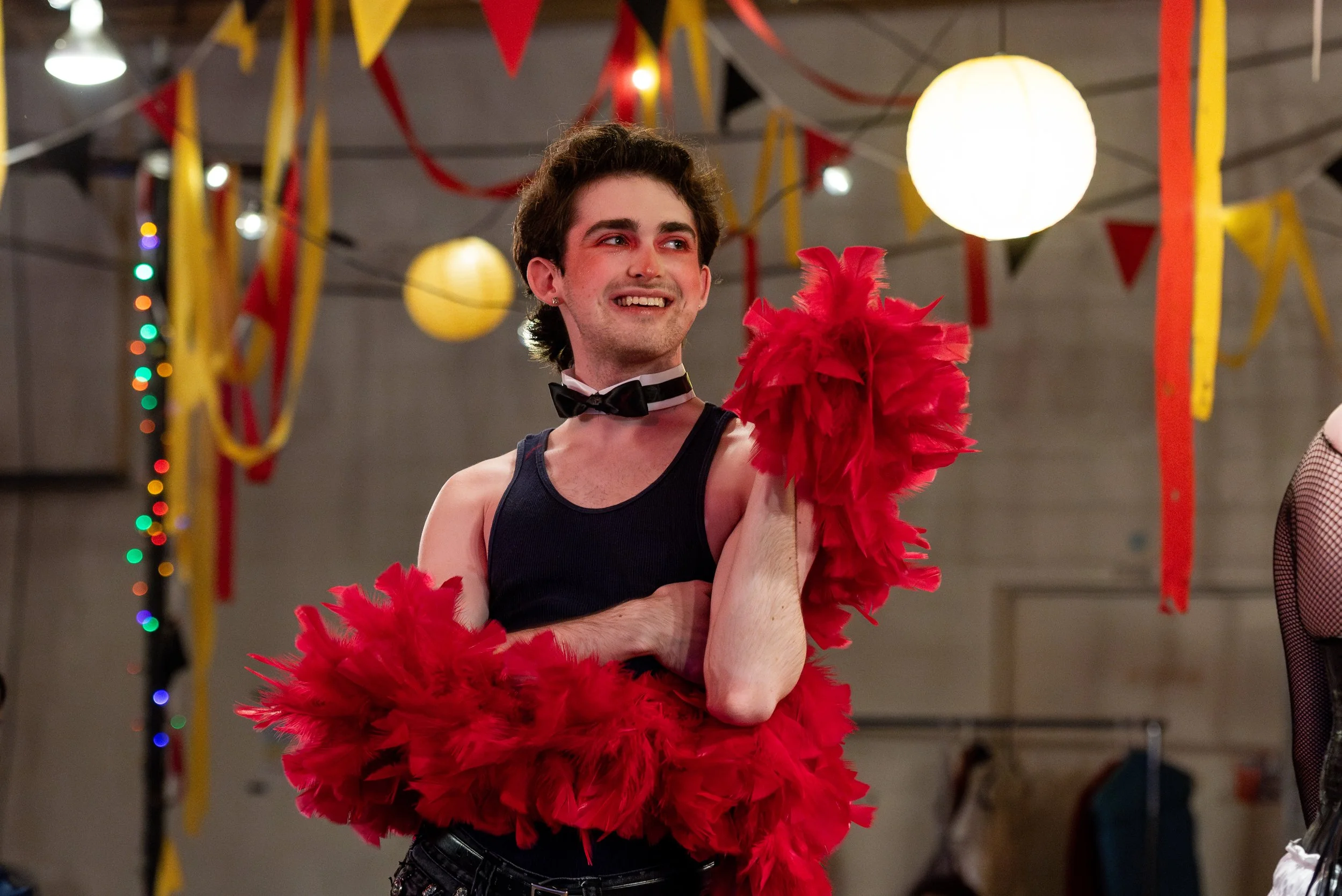 A young man dressed in a black tank top, black pants, and bow tie, smiling and holding a red feathered fan, under colorful hanging decorations and round paper lanterns at a festive party.