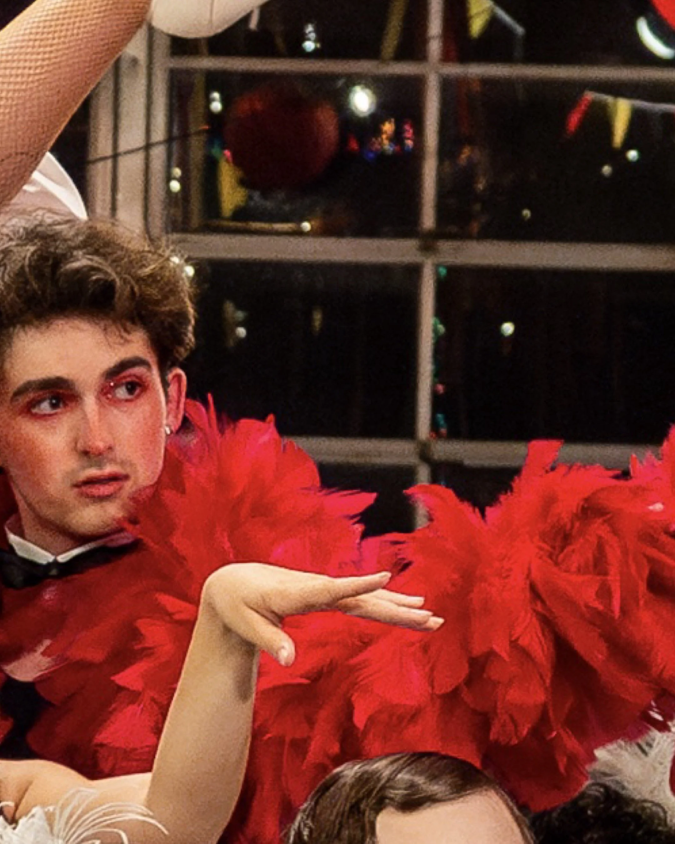 A person dressed in a red feathered costume with their hand extended, alongside another individual with curly hair and makeup, in front of a window with reflections and city lights at night.