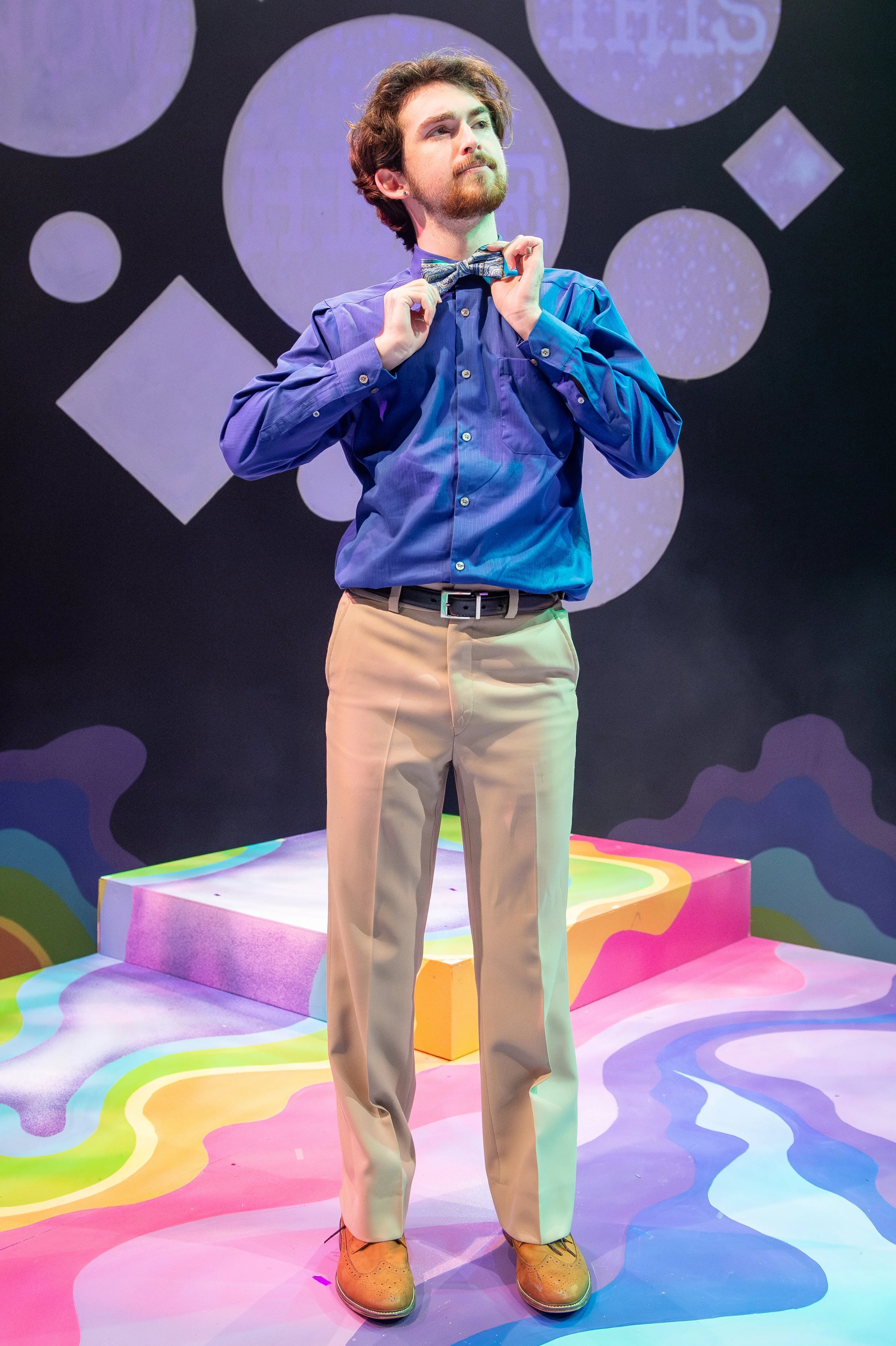 A man adjusting a bow tie on stage with a colorful, psychedelic background and abstract design patterns.