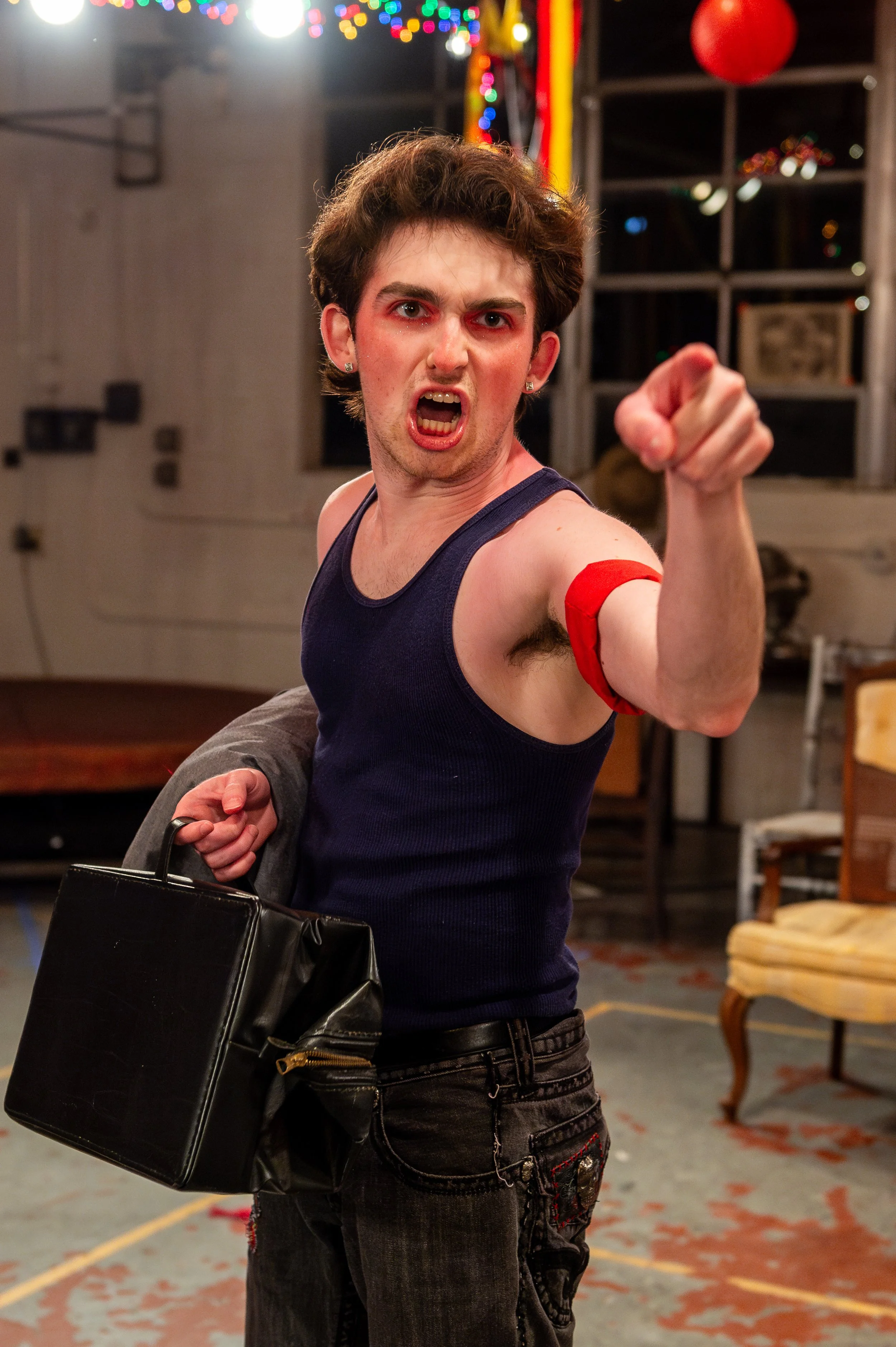 A young man with makeup and earrings, wearing a sleeveless shirt, pointing aggressively at the camera, holding a black briefcase, in a room decorated with colorful string lights and red balloons.