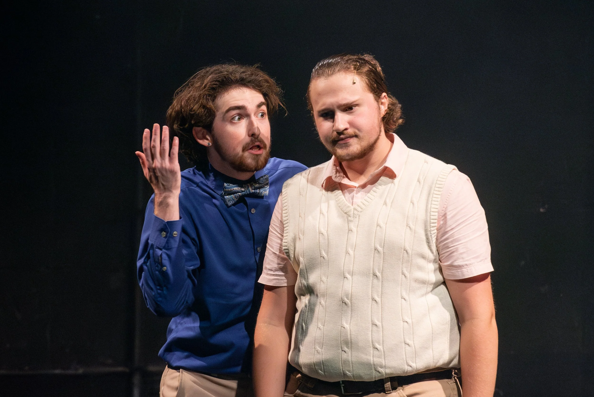 Two men performing on a stage, one wearing a blue shirt and bow tie, gesturing with his hand, the other in a light pink shirt and white sweater vest, looking at the audience with a serious expression.