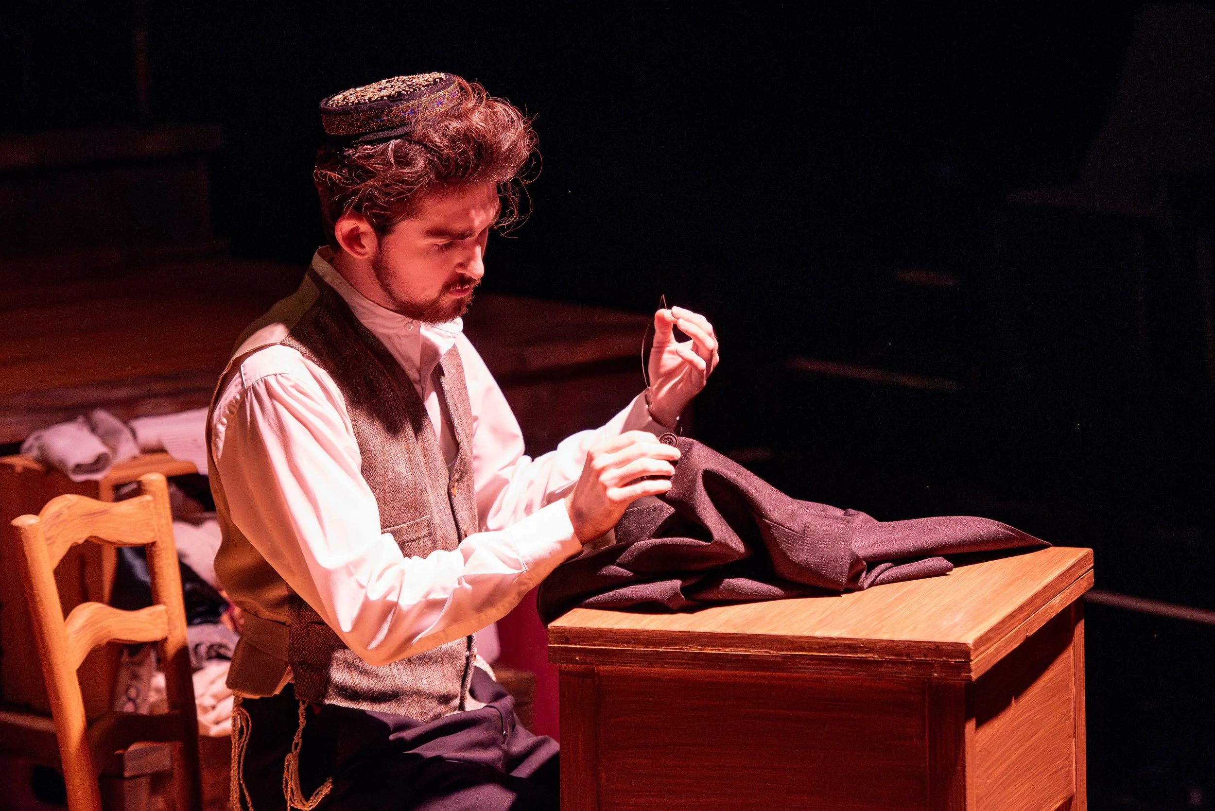 A man with curly hair and a beard, dressed in vintage clothing, sits at a wooden desk with a pile of clothes or fabric, sewing or mending a dark garment.