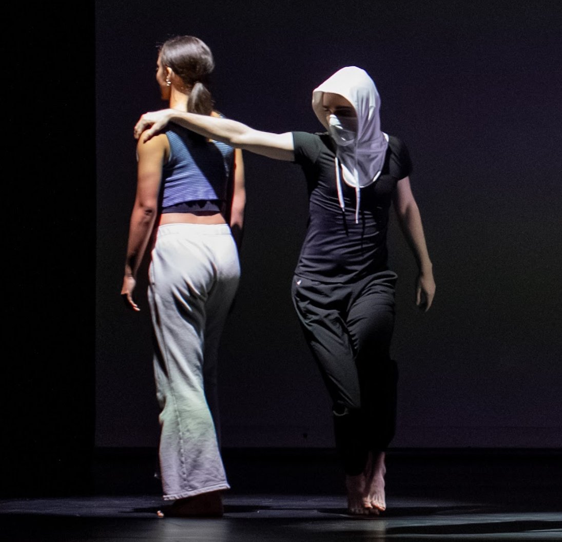Two women, one wearing a hijab and face mask, is reaching out and placing her hand on the shoulder of another woman with long hair, in a dark setting.