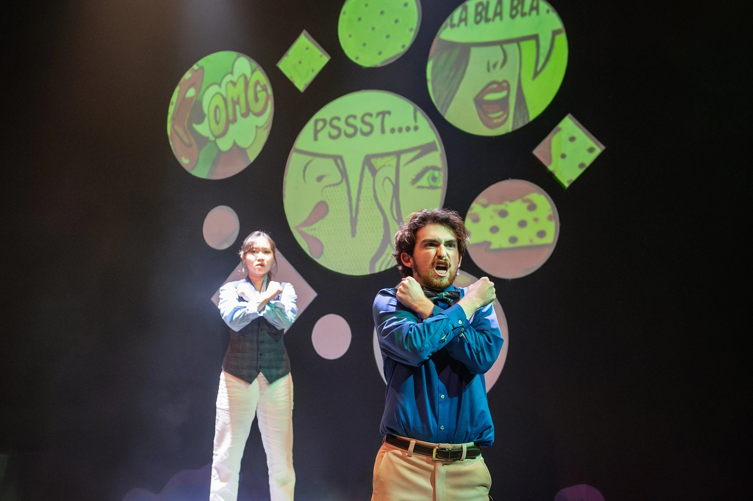 A man and woman performing on a stage with comic-style illustrations of speech bubbles behind them. The man is in the foreground with an intense expression, crossed arms, wearing a blue shirt. The woman in the background has her arms crossed and a se