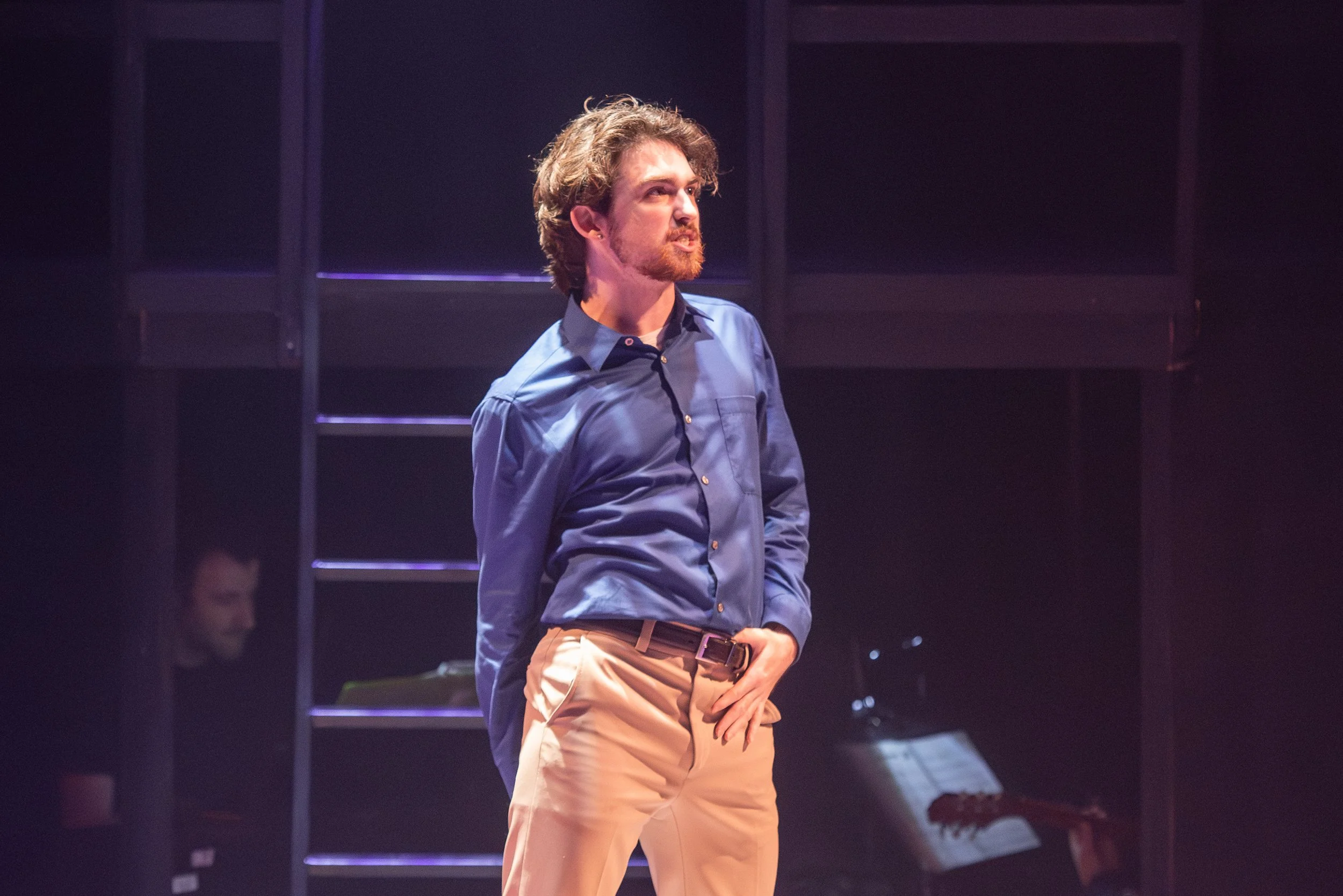 Male actor on stage during a theatrical performance, wearing a blue shirt and beige pants, with a person at a keyboard and music stand in the background.