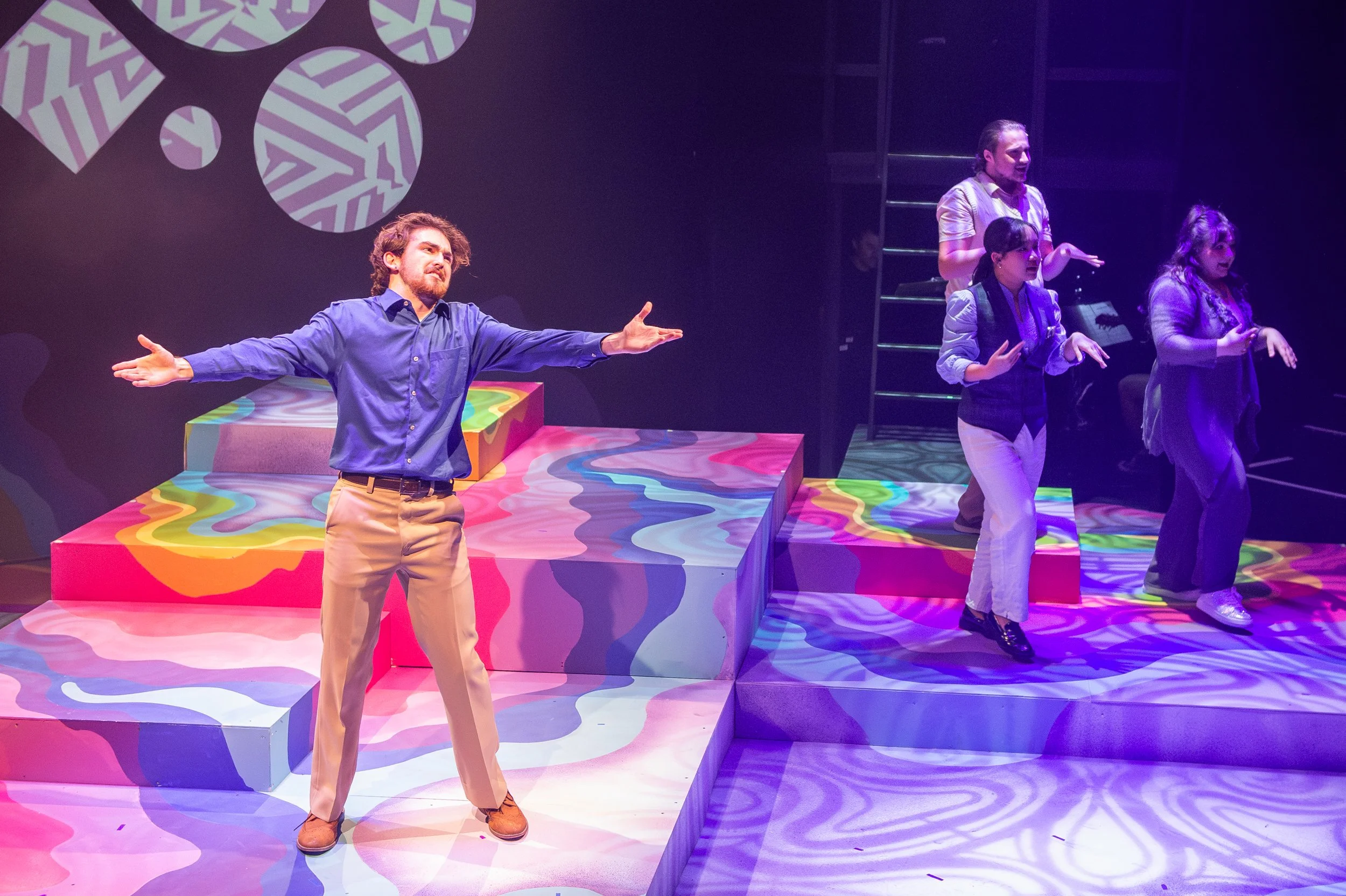 Actors performing on a colorful, abstract-set stage with geometric shapes and patterned lighting.
