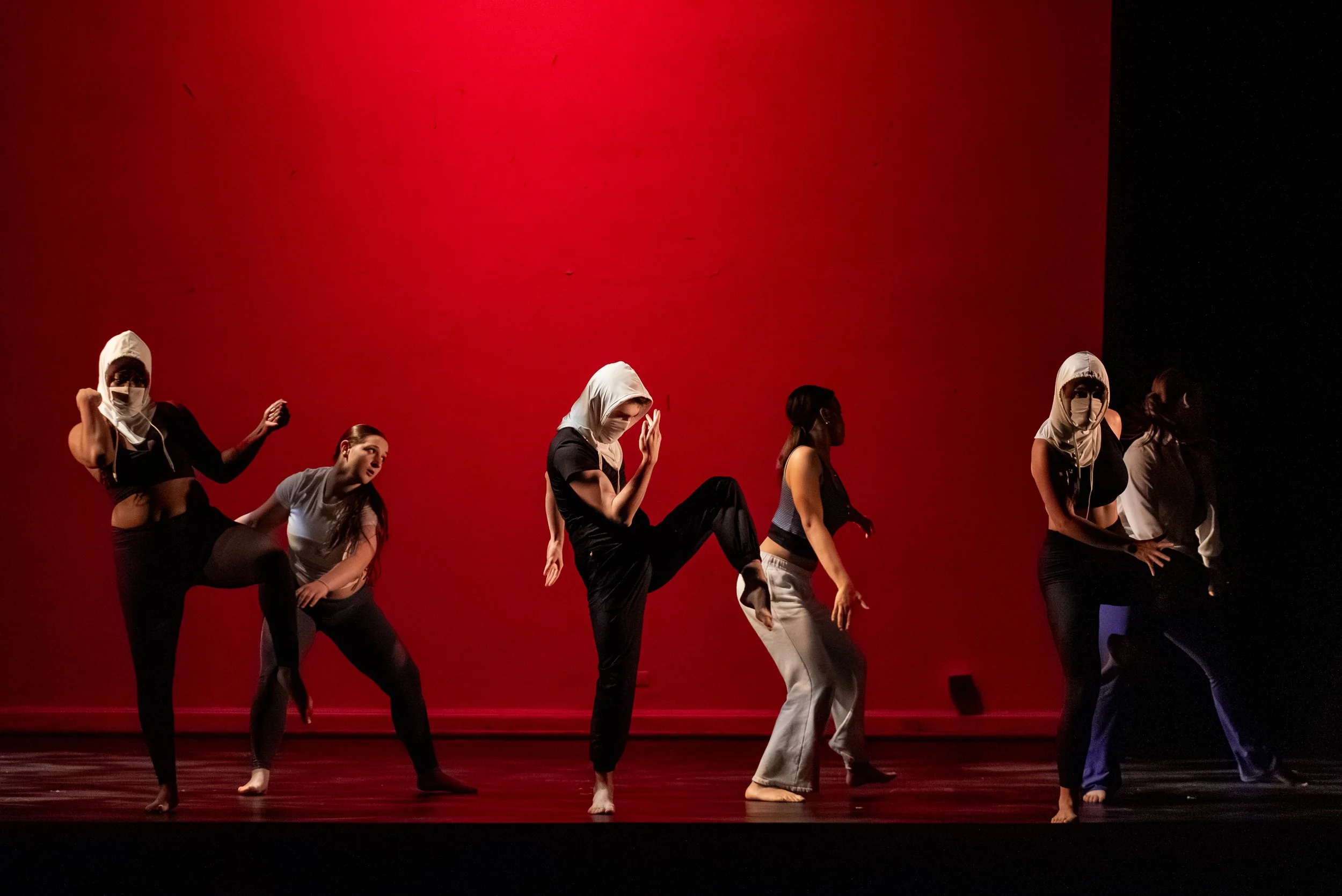 Six dancers wearing masks perform on stage with a red and black background.