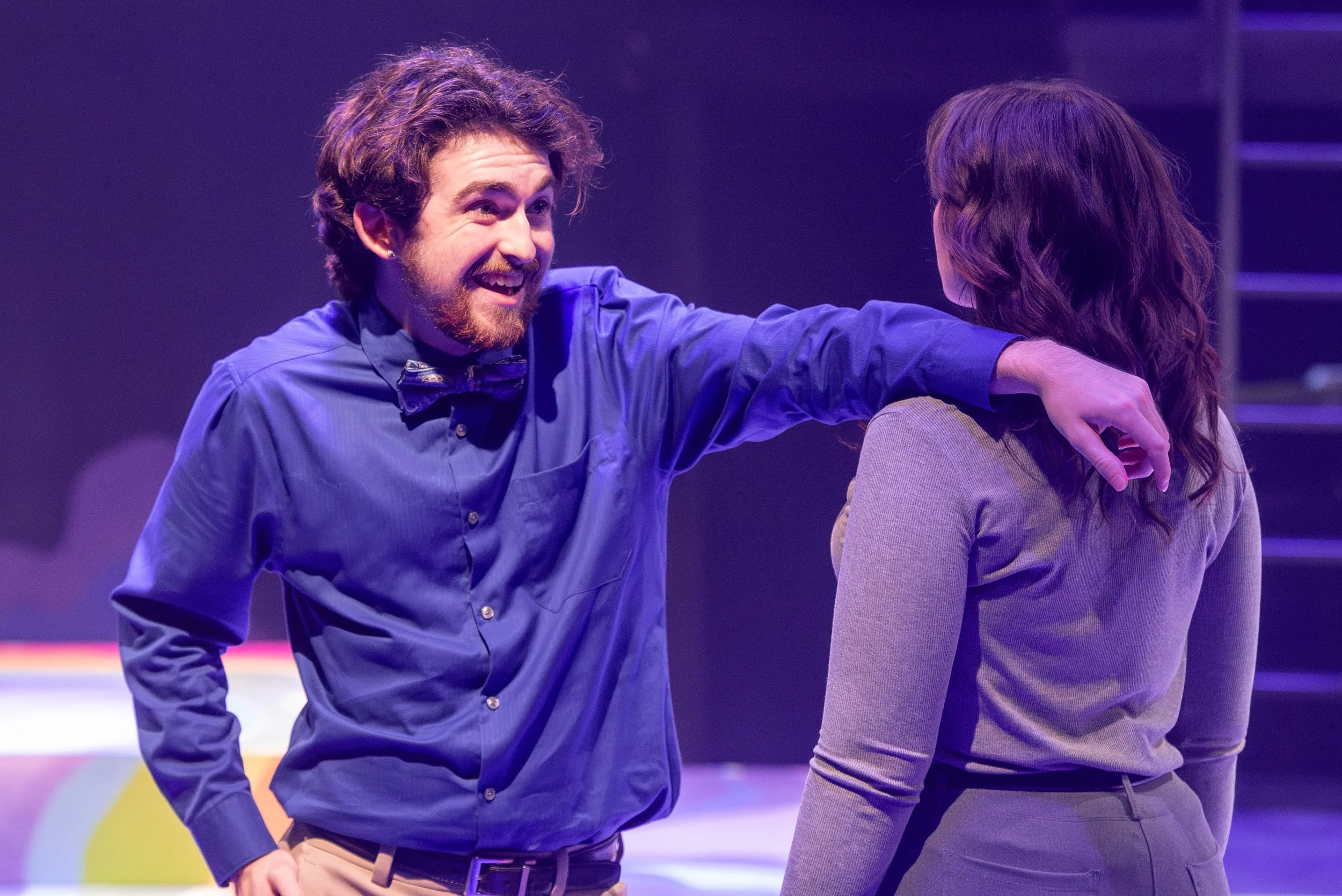 A man with curly hair and a beard wearing a blue shirt and a bow tie is smiling and talking to a woman with long wavy hair, who is wearing a gray top. The man has his arm resting on the woman's shoulder in an indoor setting with purple lighting.