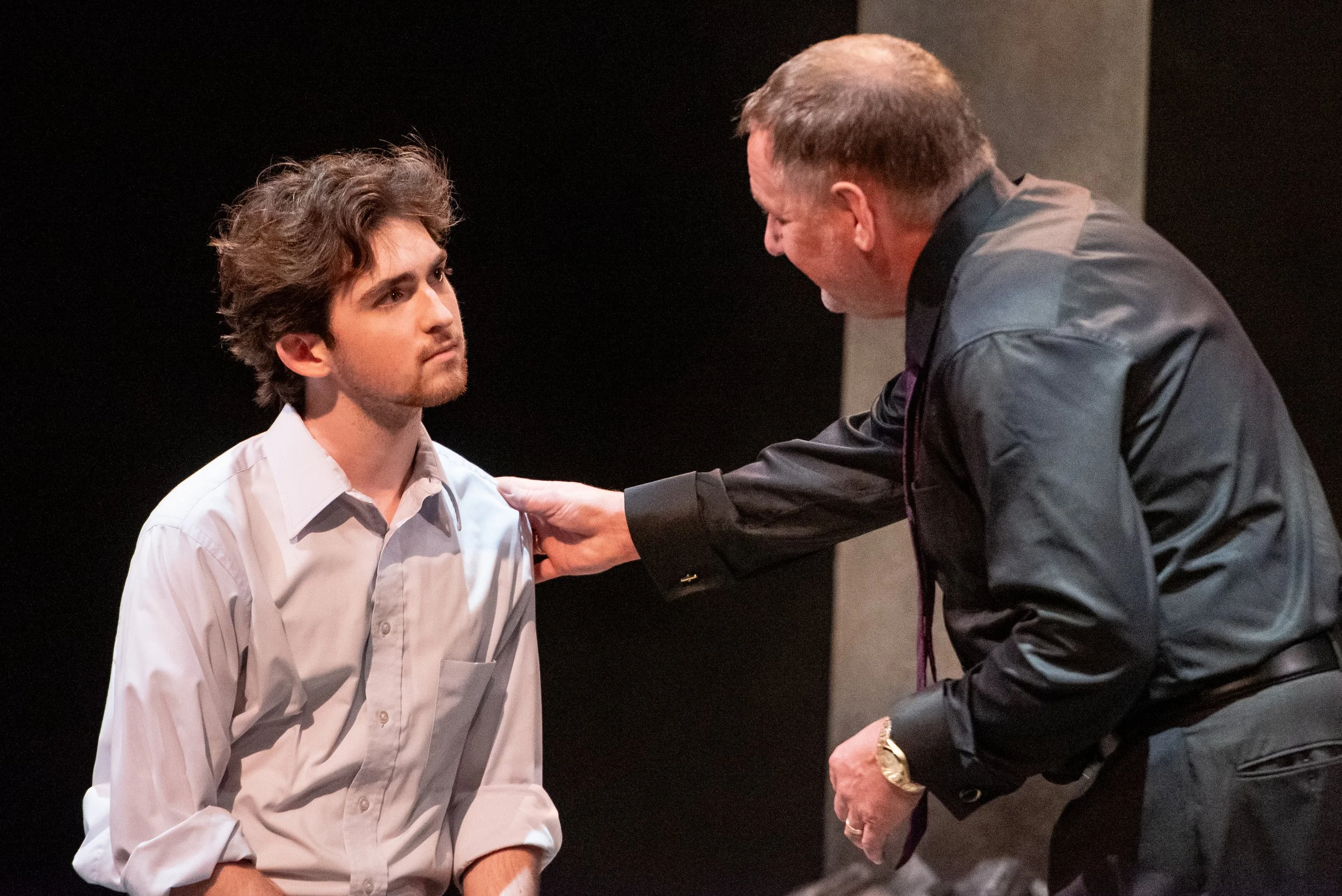 An older man in a black shirt leans toward a young man in a white shirt, placing his hand on the young man's shoulder and speaking to him, with a serious expression. The background is dark and blurred.