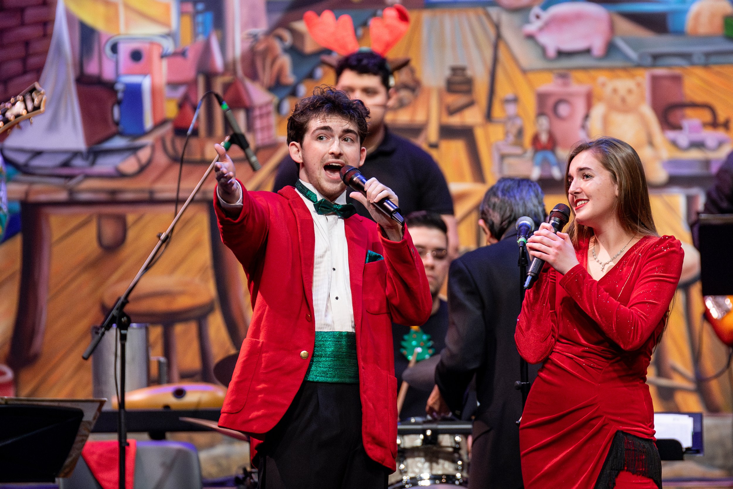 Two performers, a young man in a red jacket and a young woman in a red dress, singing into microphones on stage during a festive event with a painted backdrop and band members in the background.