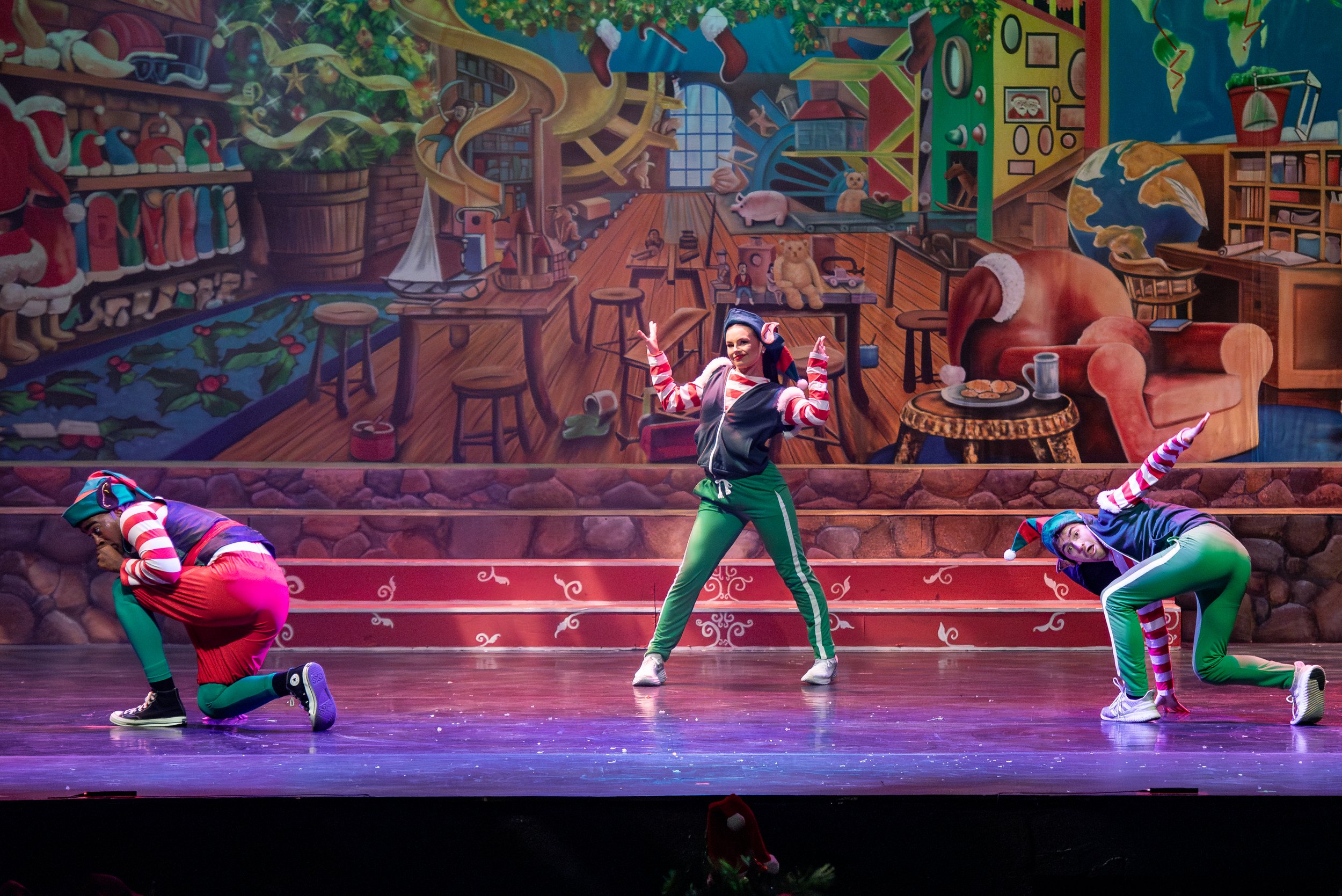 Three dancers dressed in holiday-themed costumes performing on stage with a festive, colorful Christmas-themed backdrop.