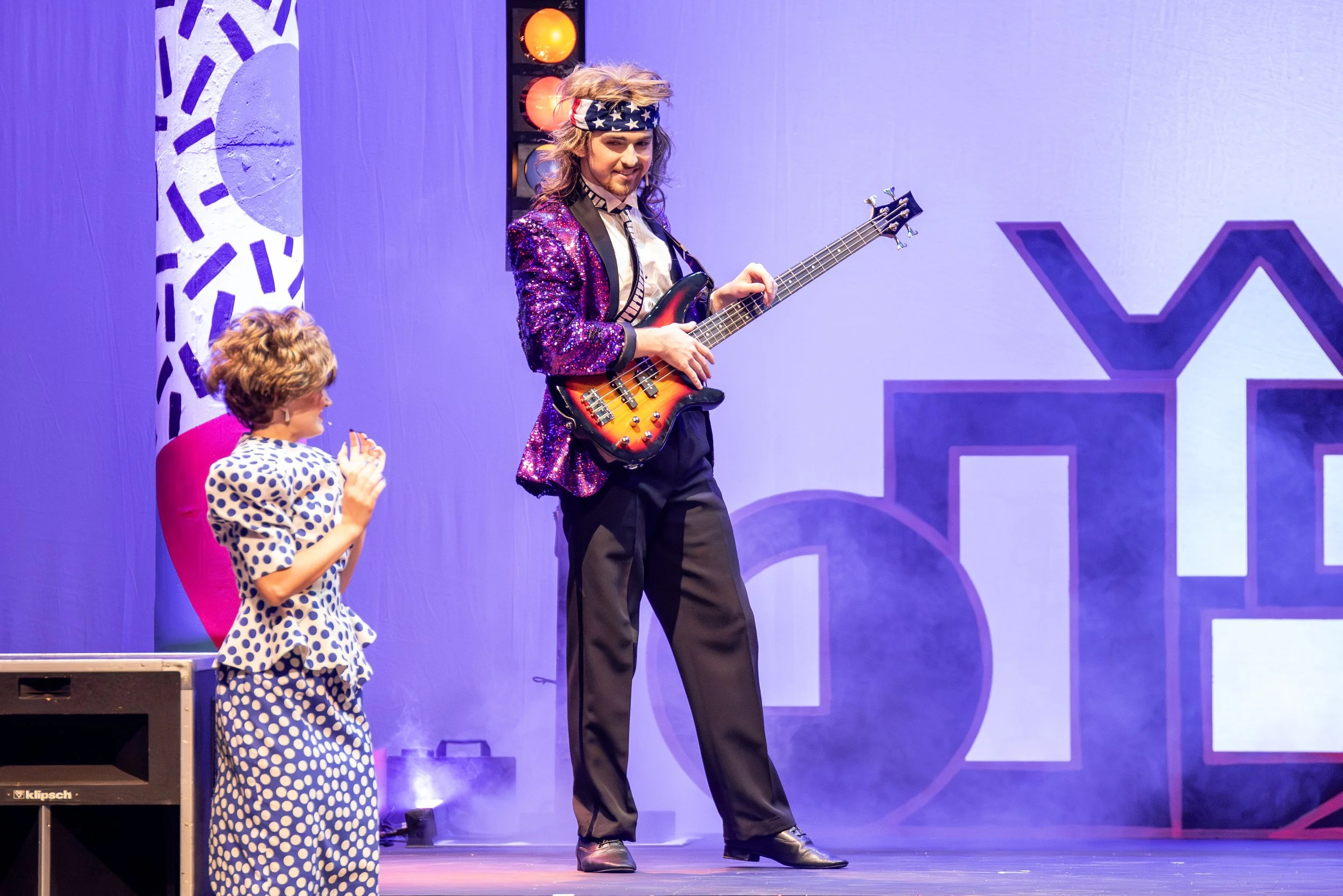 A magician with long hair, wearing a sparkly purple jacket and a star-spangled headband, plays an electric guitar on stage. A young girl in polka-dot pajamas watches and claps beside him.