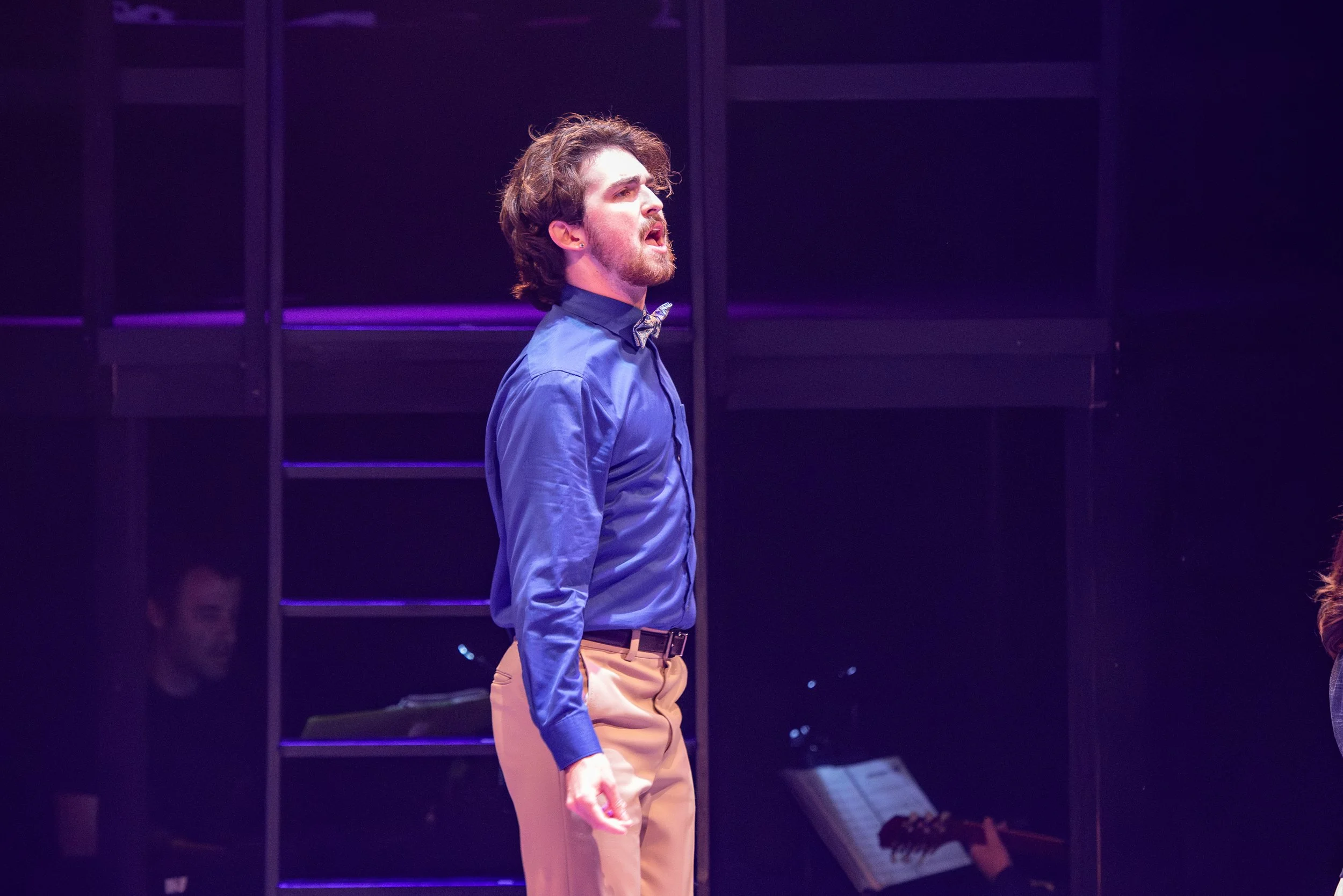 A man singing on stage wearing a blue shirt, beige pants, and a bow tie, with purple stage lighting.