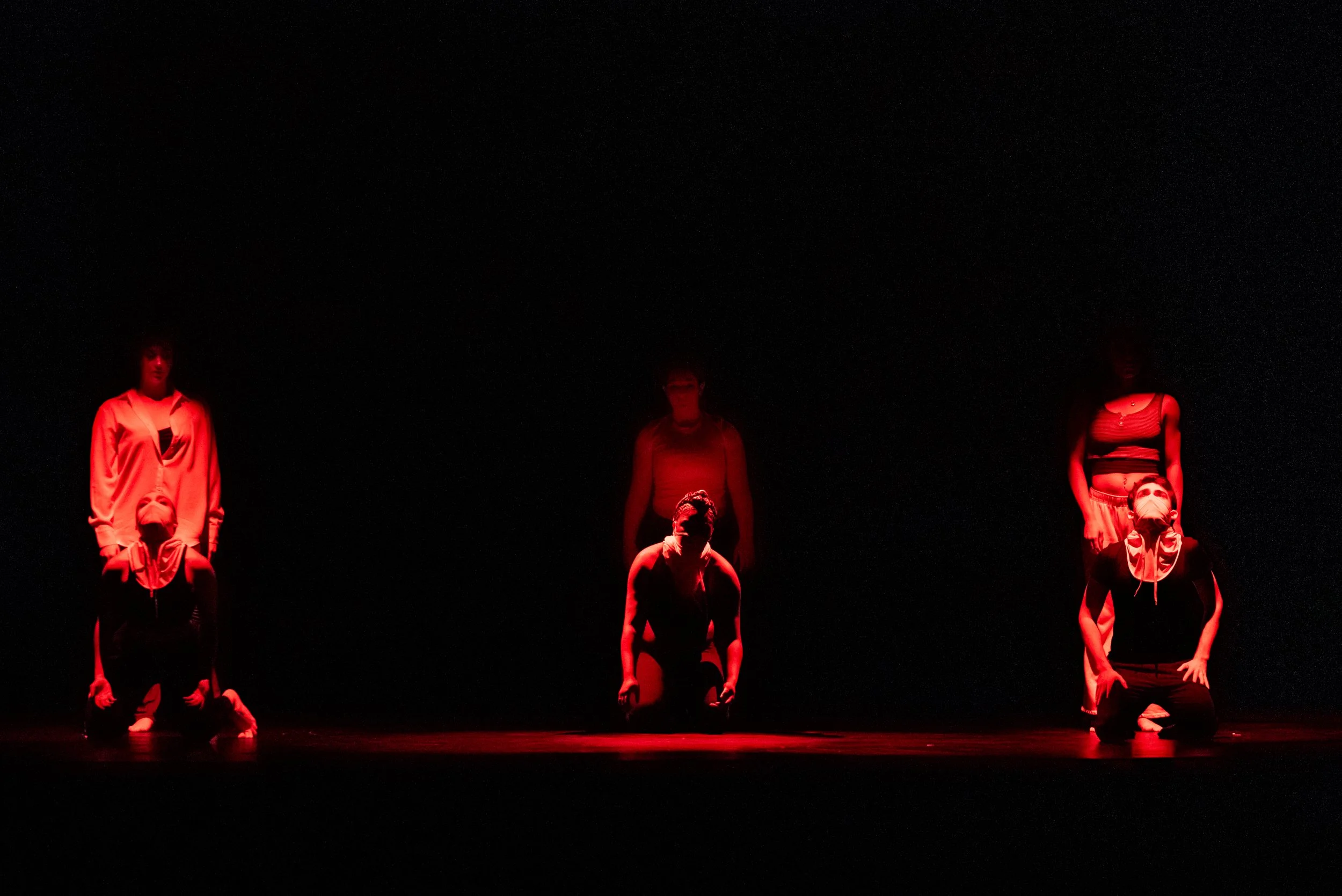 Six dancers performing on stage under red lighting against a black background, with some kneeling and others standing.
