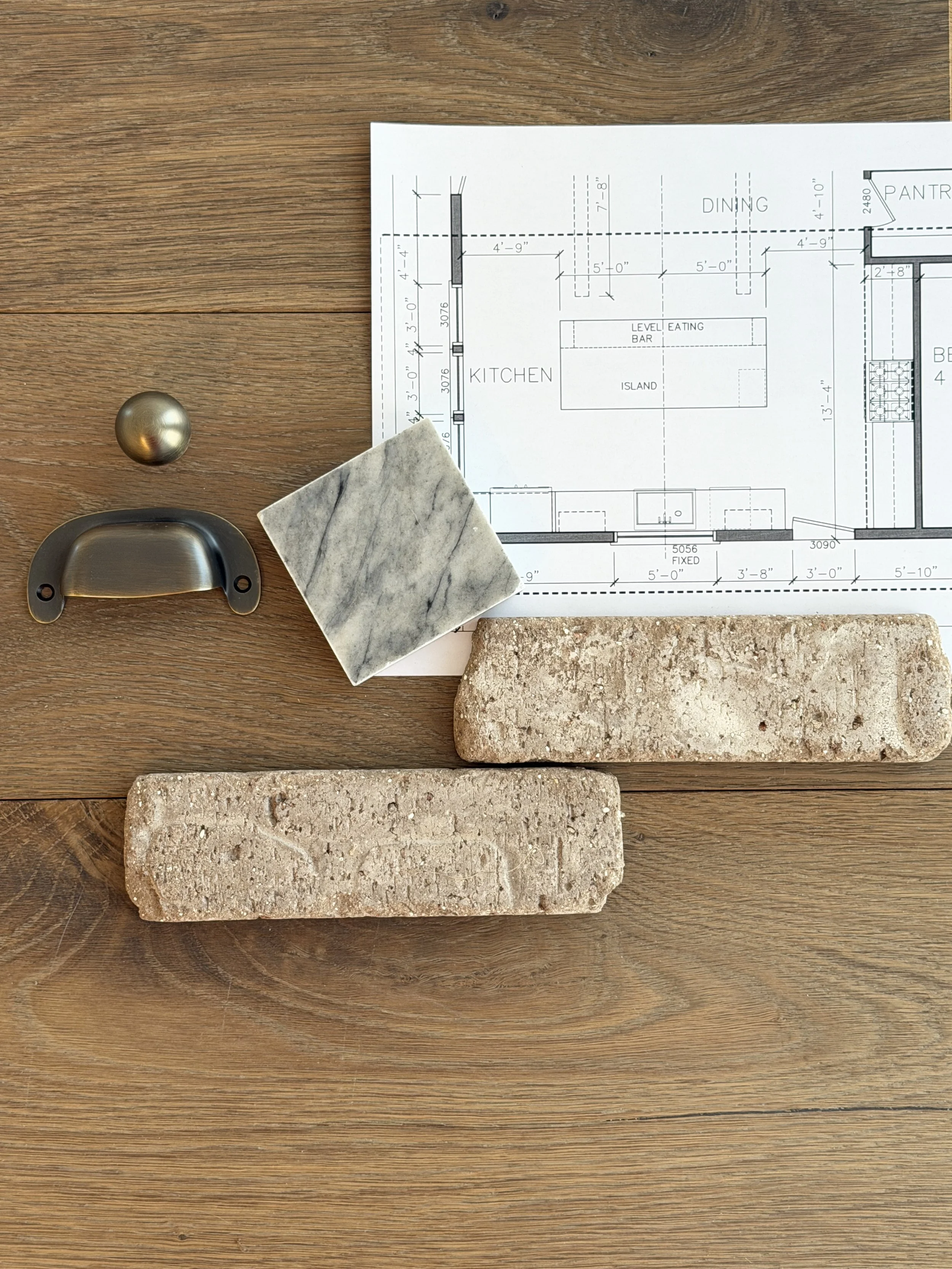 A wooden table with architectural plans, a marble square tile, a metal handle, and a brick sample placed on it.