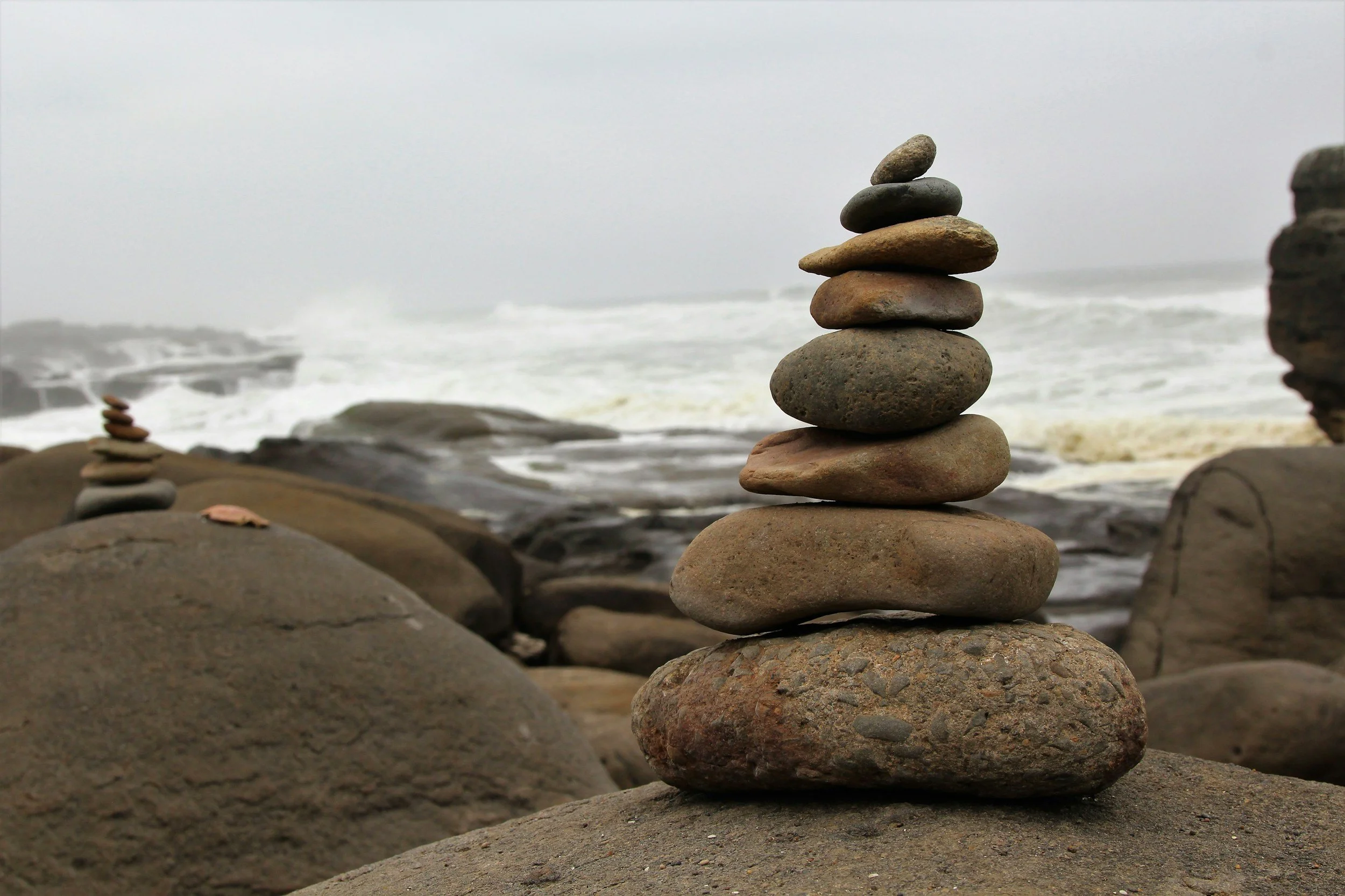 Stacked rocks balanced on driftwood on a beach, with blurred trees and ocean in the background during sunset or dusk.