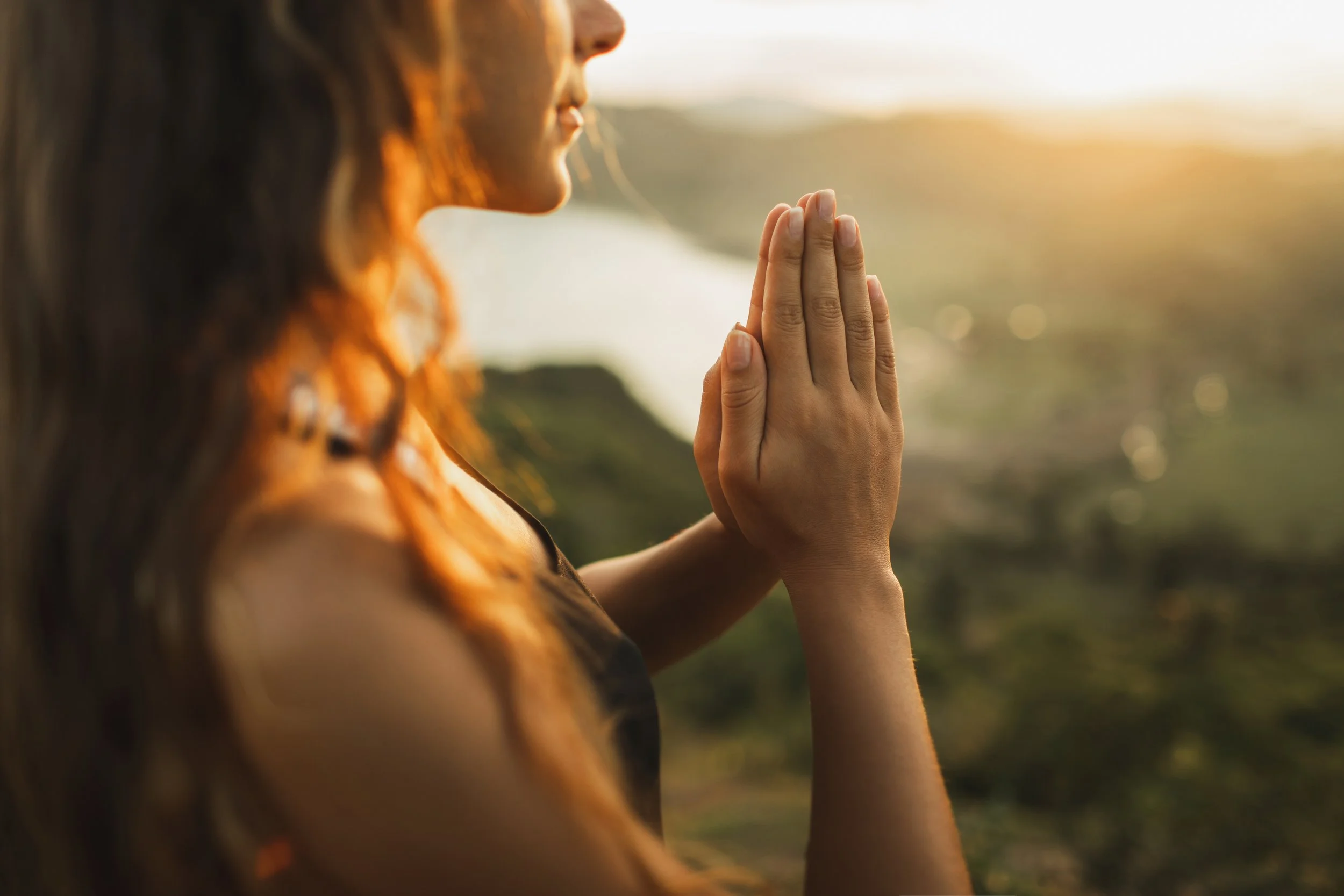 A woman with red hair practicing yoga outdoors at sunrise, with her hands in prayer position and her face turned towards the light.
