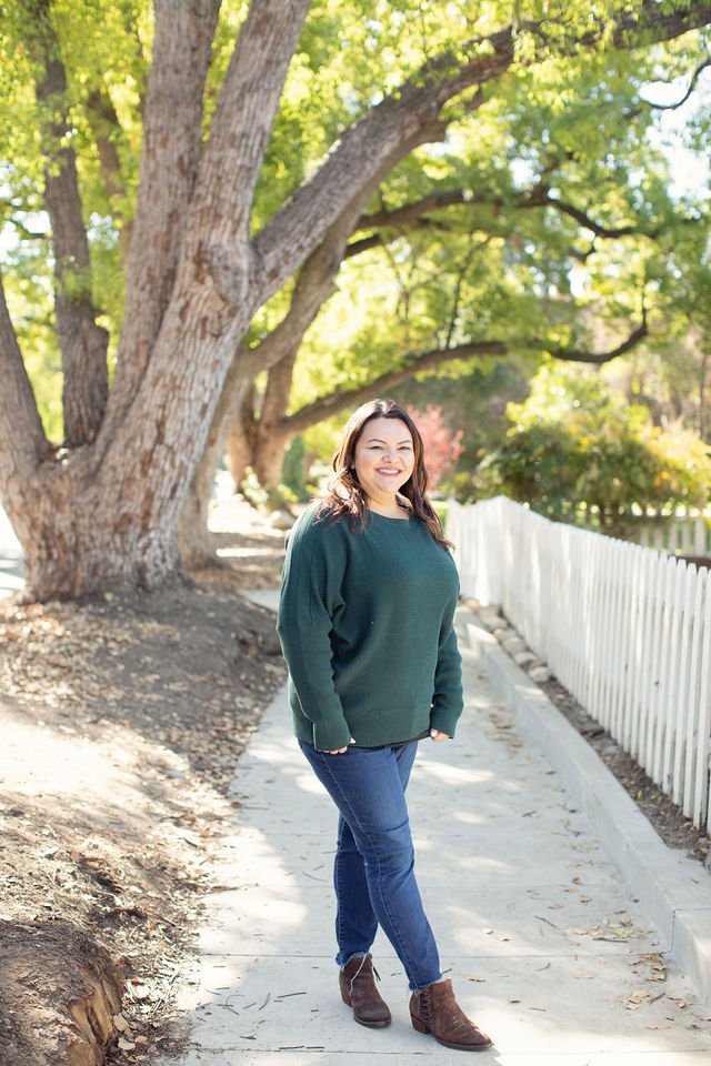 Smiling woman walking outdoors on a sidewalk with trees and a white fence in the background.