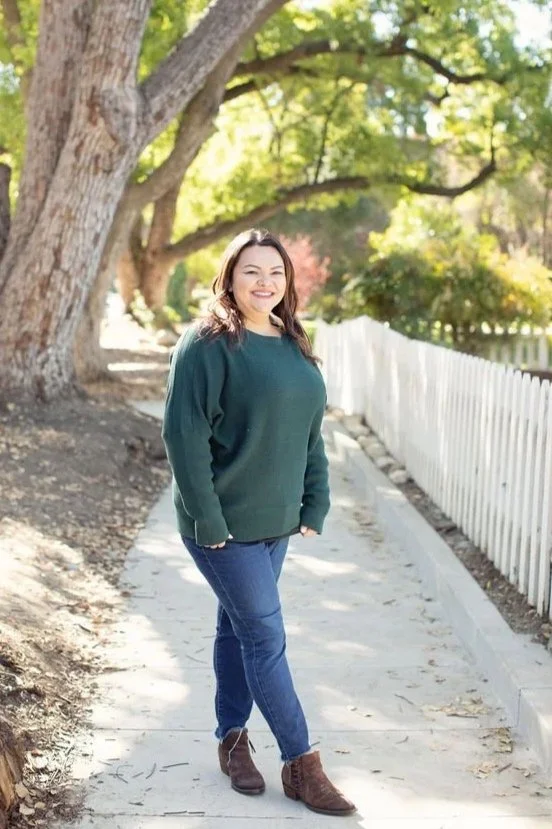 A woman with shoulder-length brown hair smiling, wearing a dark green sweater, blue jeans, and brown ankle boots, standing on a sunlit sidewalk next to a white picket fence and large trees with green leaves.