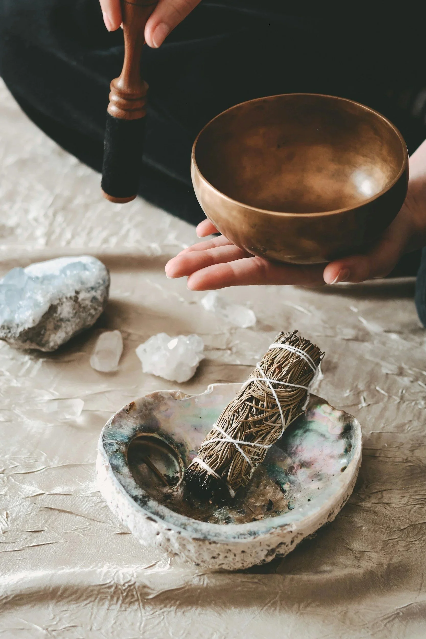 A person holding a singing bowl with a mallet, surrounded by rocks, crystals, and a bundle of sage or herbs on a textured surface.