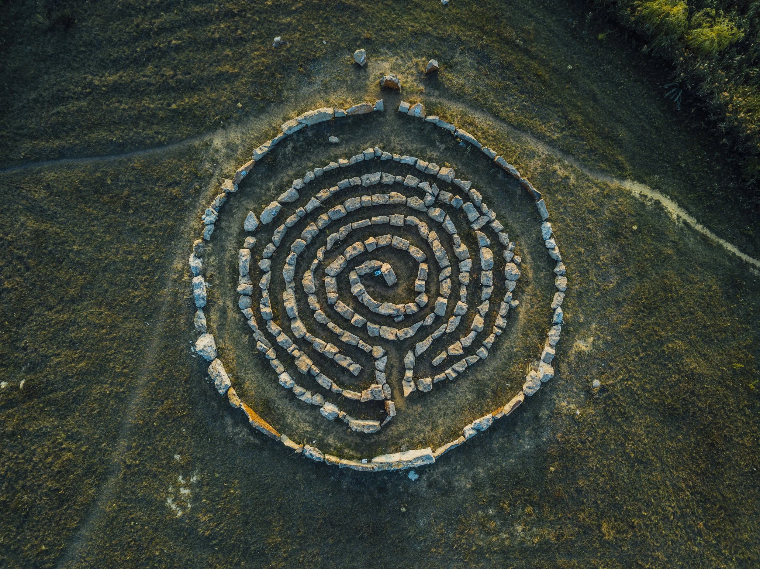 An aerial view of a stone labyrinth in a grassy field, with walking paths leading away from it.