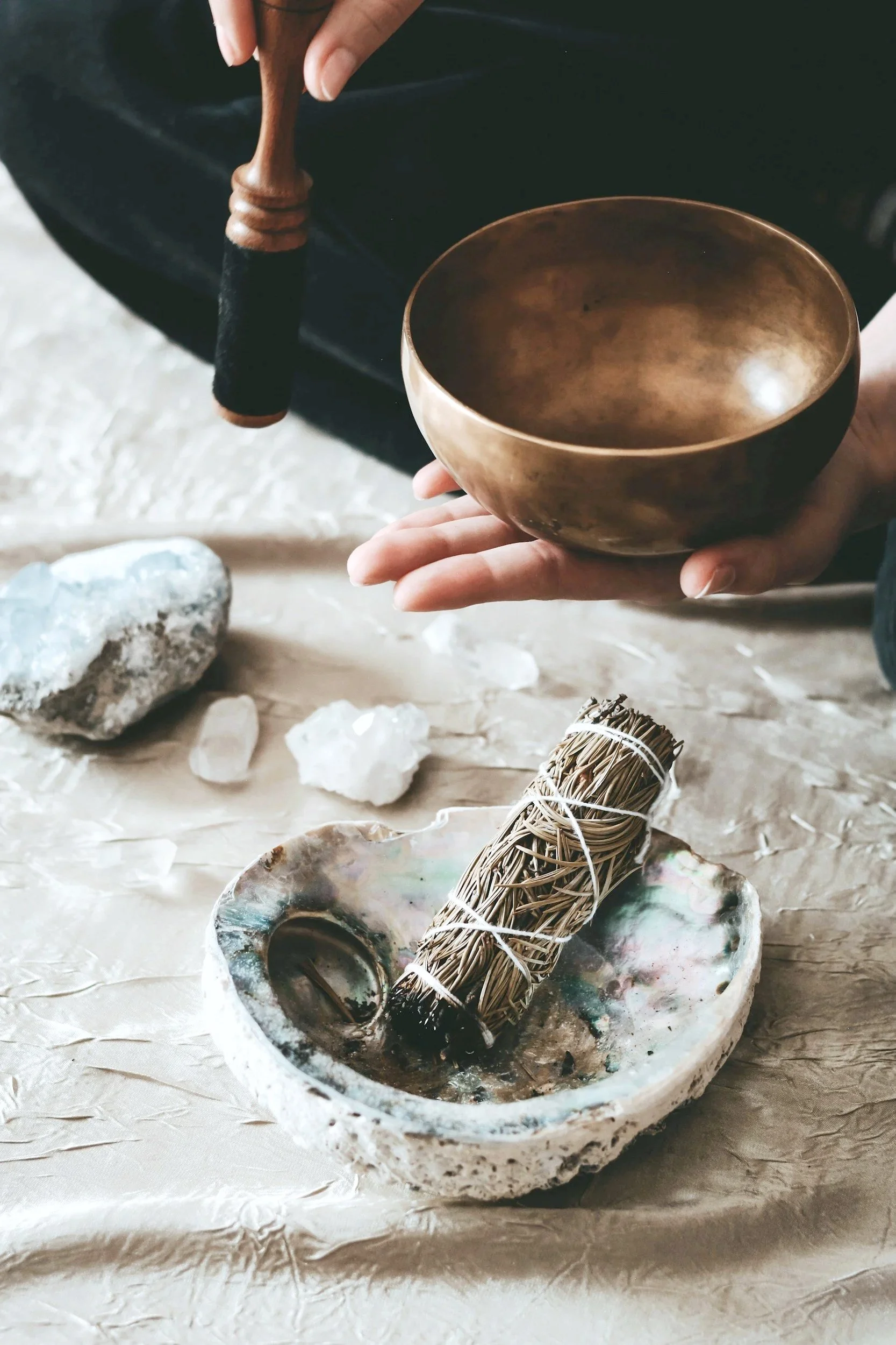 Person holding a brass singing bowl with a wooden striker in their other hand, with crystals and a bundle of herbs or sage on a textured surface.
