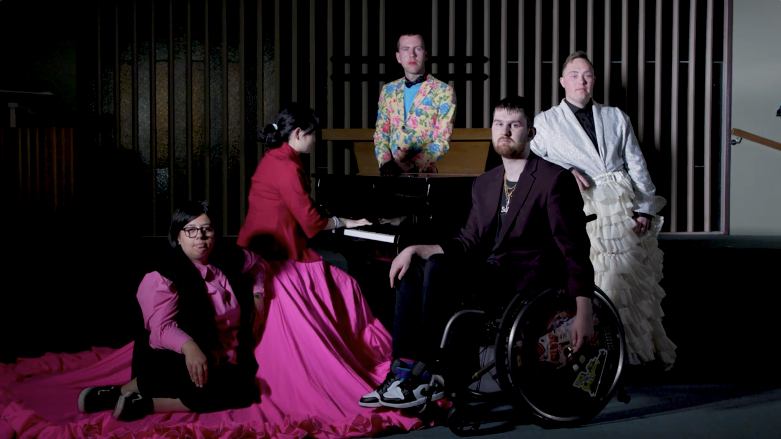 Five actors are posed around a grand piano. They are dressed in colourful fancy clothes and have serious expressions on their faces.