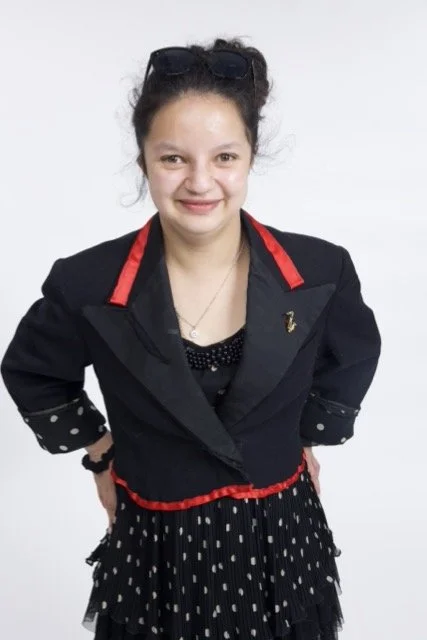 Azarah stands with her hands on the back of her hips. She is wearing a black jacket with red trim. She is smiling. Her hair is pulled back into a bun.