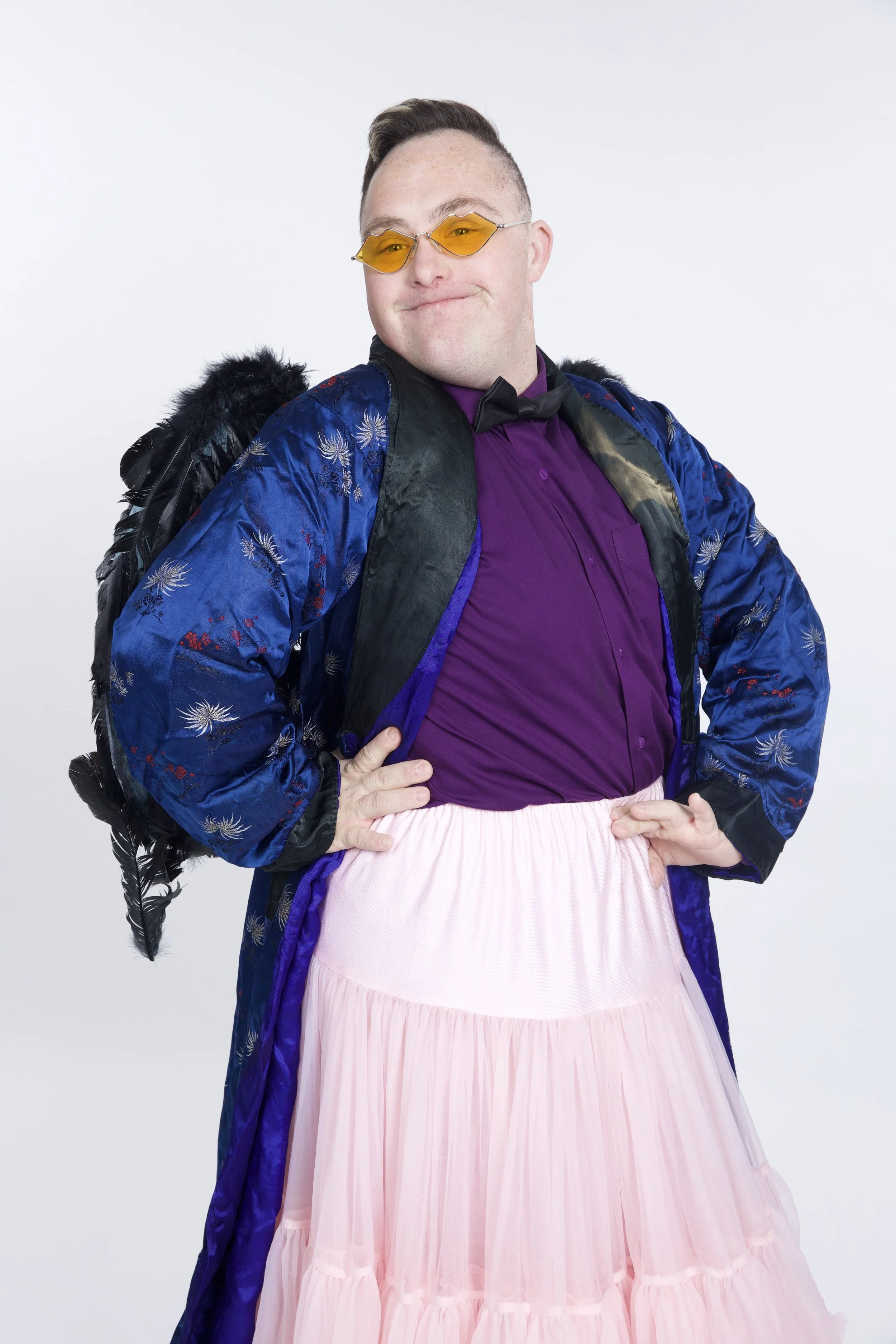 Callum has short brown hair, and is wearing a blue jacket with a purple top and a pink skirt. He is wearing yellow sunglasses, and is smiling, with his hands on his hips.