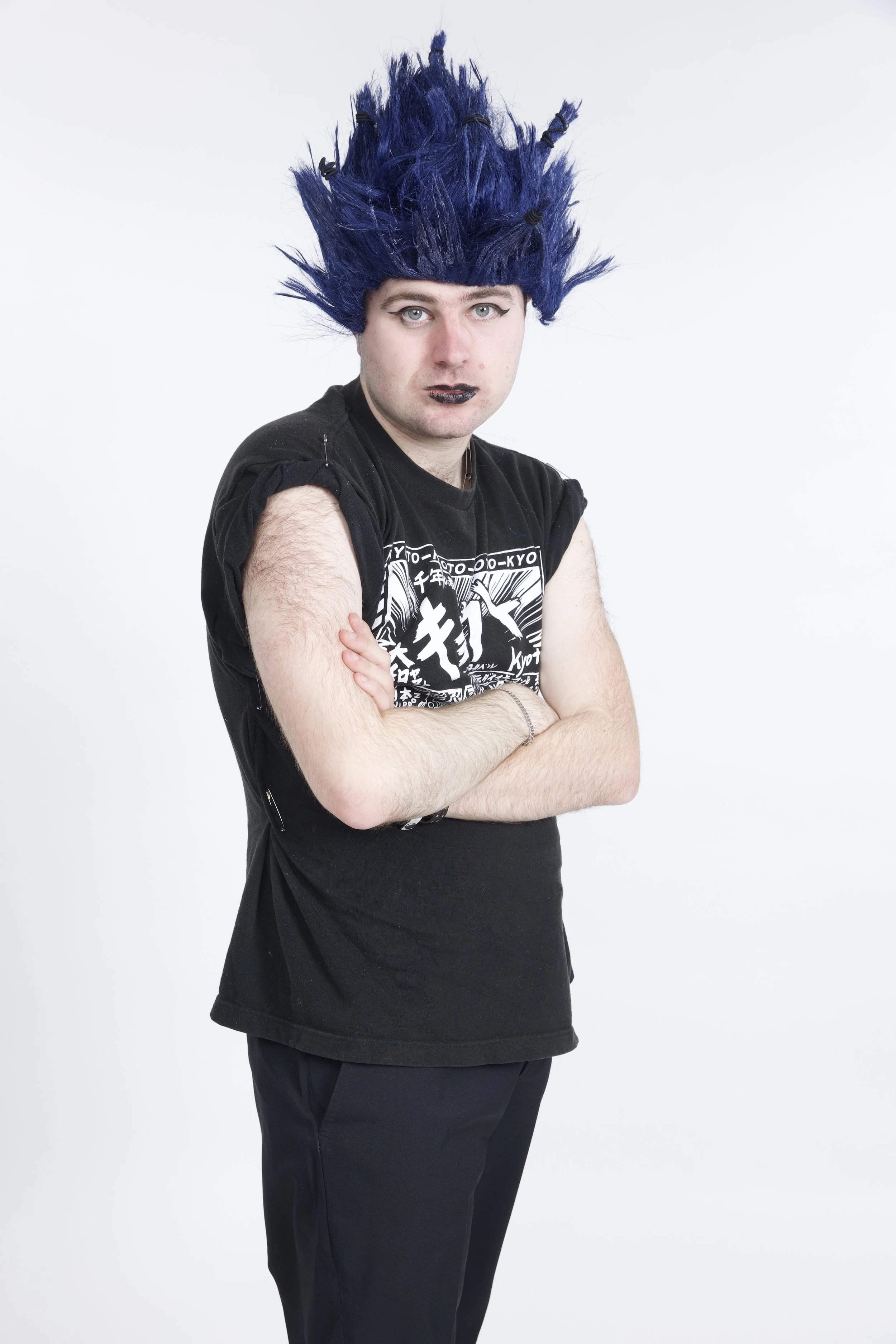 Leo stands with his arms folded. He is wearing a blue wig which is all spiked up in a punk style. He has black eyeliner and black lipstick on.