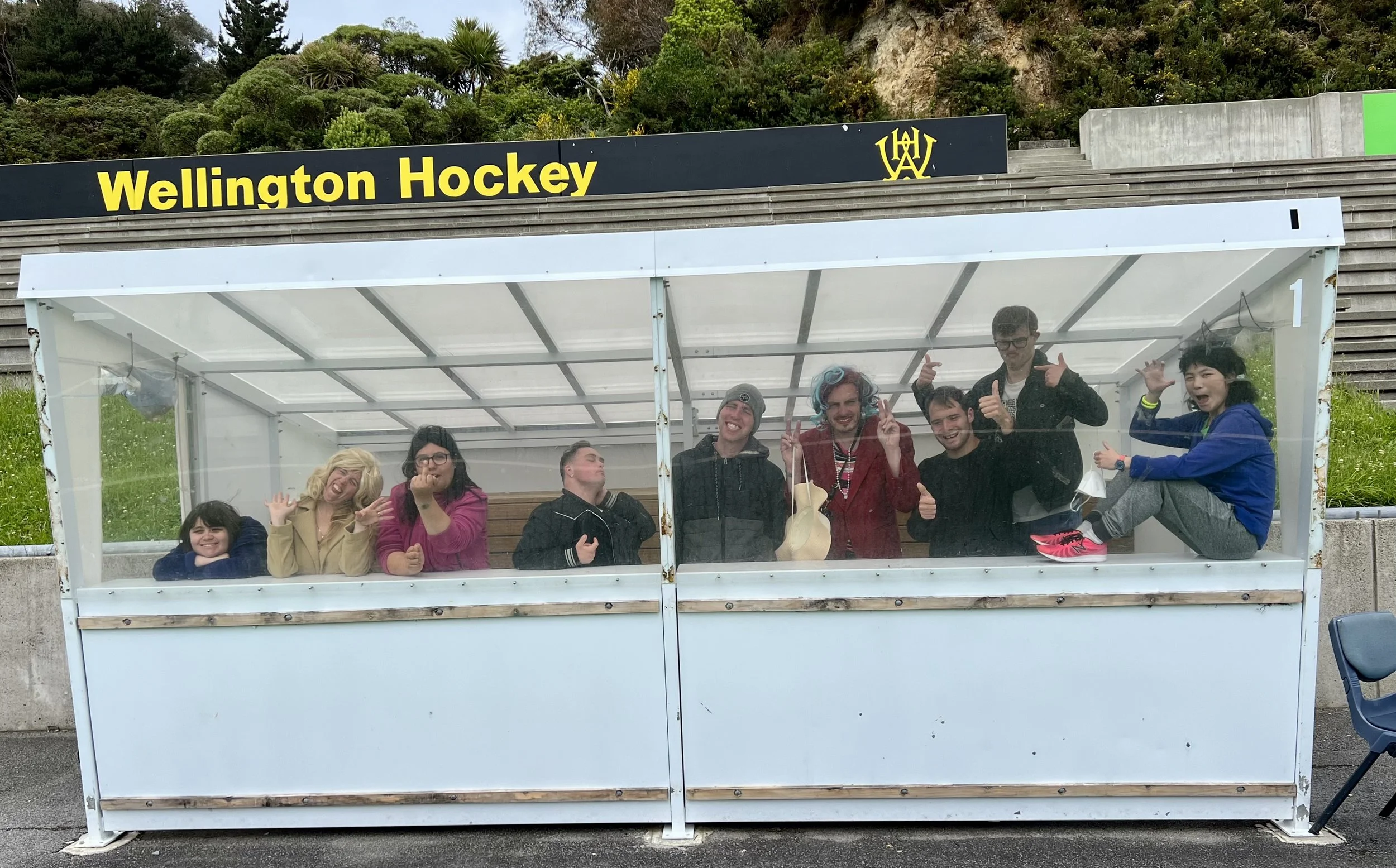 9 actors stand and sit posed inside a hockey viewing box. There is a sign at the top that reads "Wellington Hockey". They are all looking at the camera and smiling.