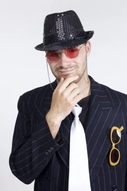 Jeremy is wearing a suit and tie, with a sparkly fedora and red-tinted sunglasses. He has his hand up on his chin in a pensive fashion.