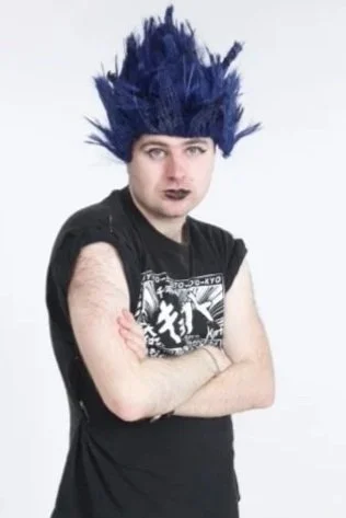 Leo stands with his arms folded. He is wearing a blue wig that is all spiked up in a punk style, and a short sleeved black shirt with a white print on the front. He has dark lipstick on.